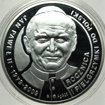 Srebrna moneta kolekcjonerska upamiętniająca Jana Pawła II, z wygrawerowanym wizerunkiem papieża i napisem 'Rocznica I Pielgrzymki Do Polski 1979-2009', umieszczona w przezroczystym etui.