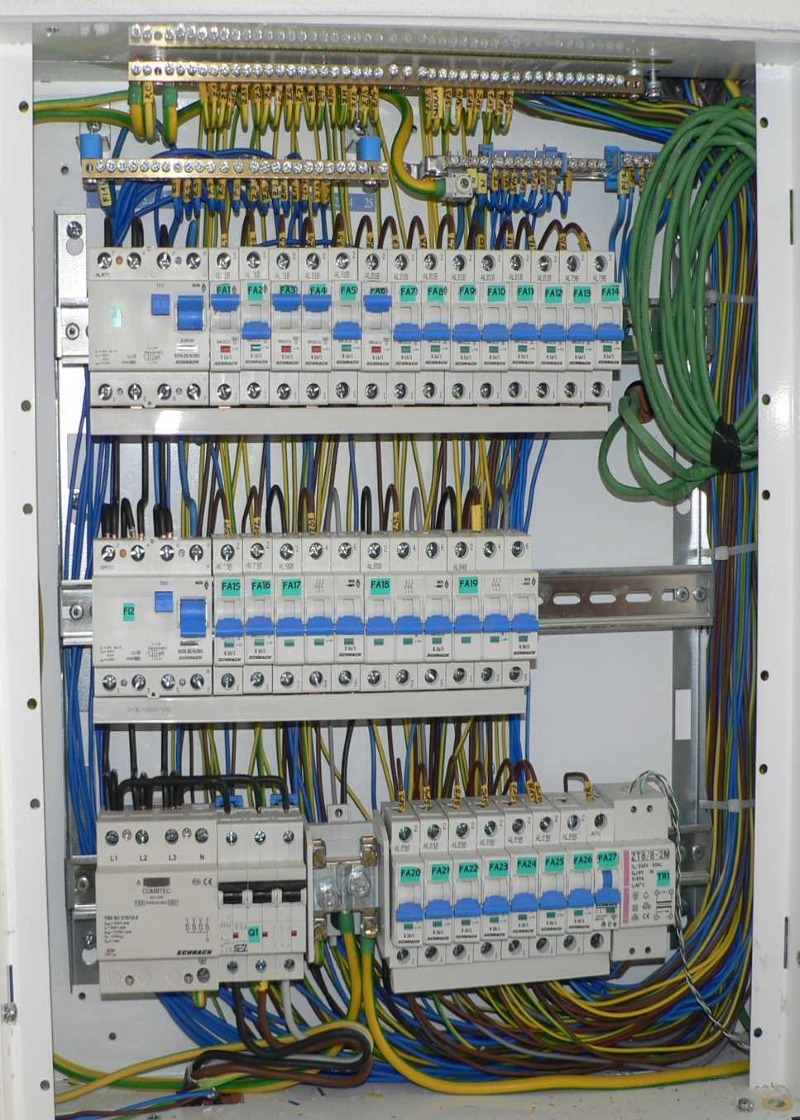 Otwarta skrzynka elektryczna z zainstalowanymi bezpiecznikami, okablowaniem w kolorach niebieskim, żółtym, zielonym i brązowym, oraz listwami zaciskowymi.