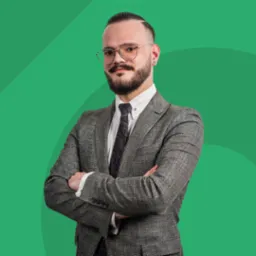 Portret mężczyzny w okularach i tweedowym garniturze, z brodą i wąsami, stojącego na tle zielonego studia z założonymi rękami.