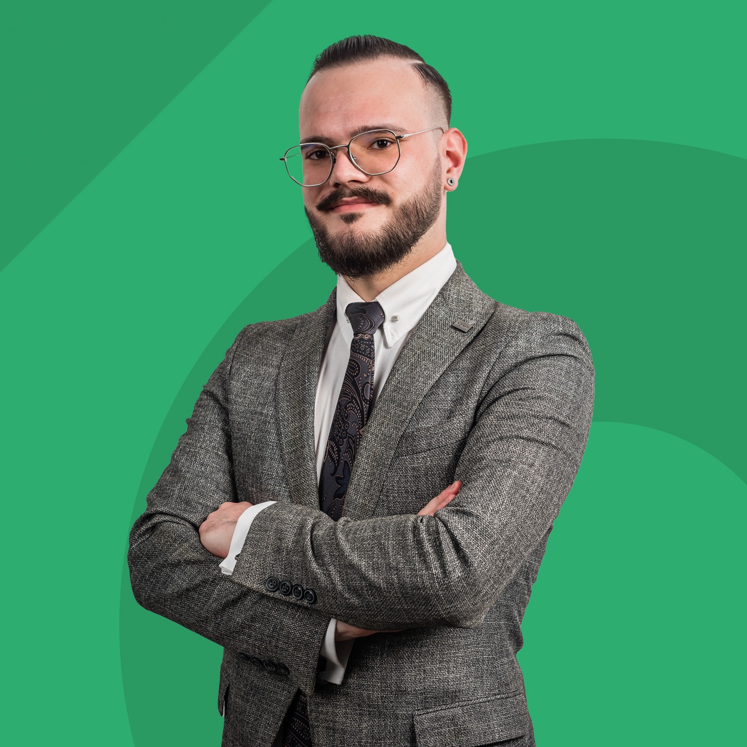 Portret mężczyzny w okularach i tweedowym garniturze, z brodą i wąsami, stojącego na tle zielonego studia z założonymi rękami.