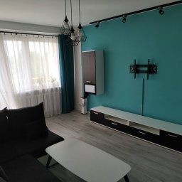 Colorful - Turkusowa ściana w salonie z zamontowanym uchwytem TV, nowoczesne oświetlenie sufitowe i minimalistyczne meble w odcieniach bieli i czerni.