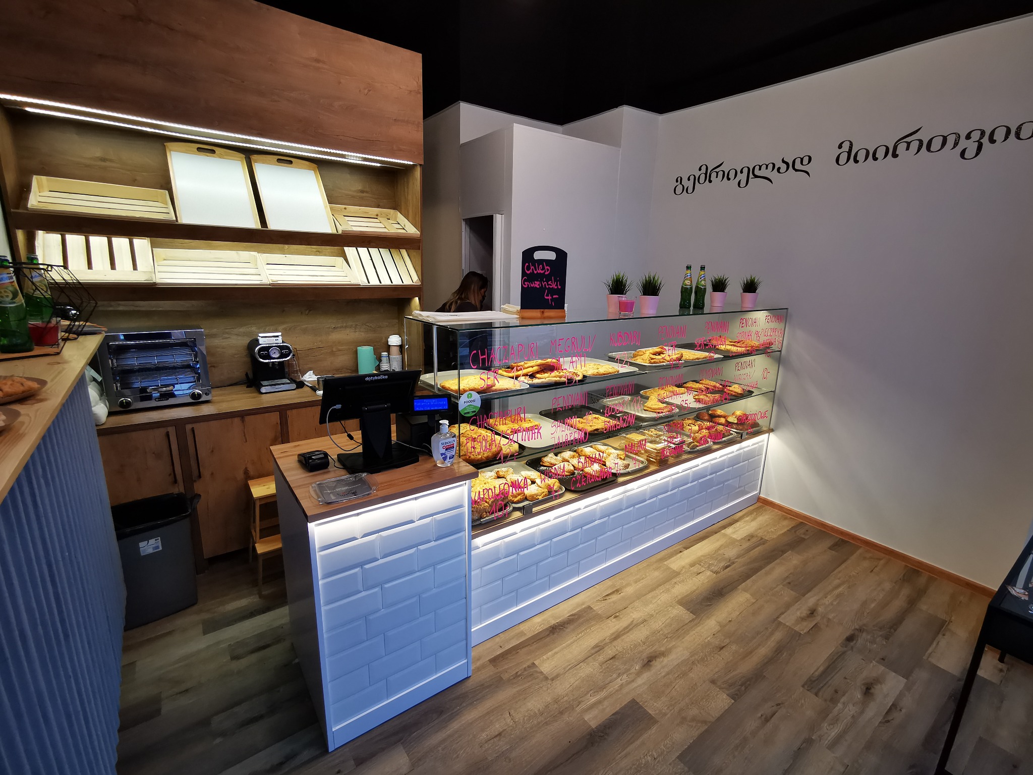 Wnętrze piekarni z drewnianymi półkami, przeszkloną ladą chłodniczą pełną gruzińskich wypieków, kasą fiskalną i białymi płytkami na ścianach lady.