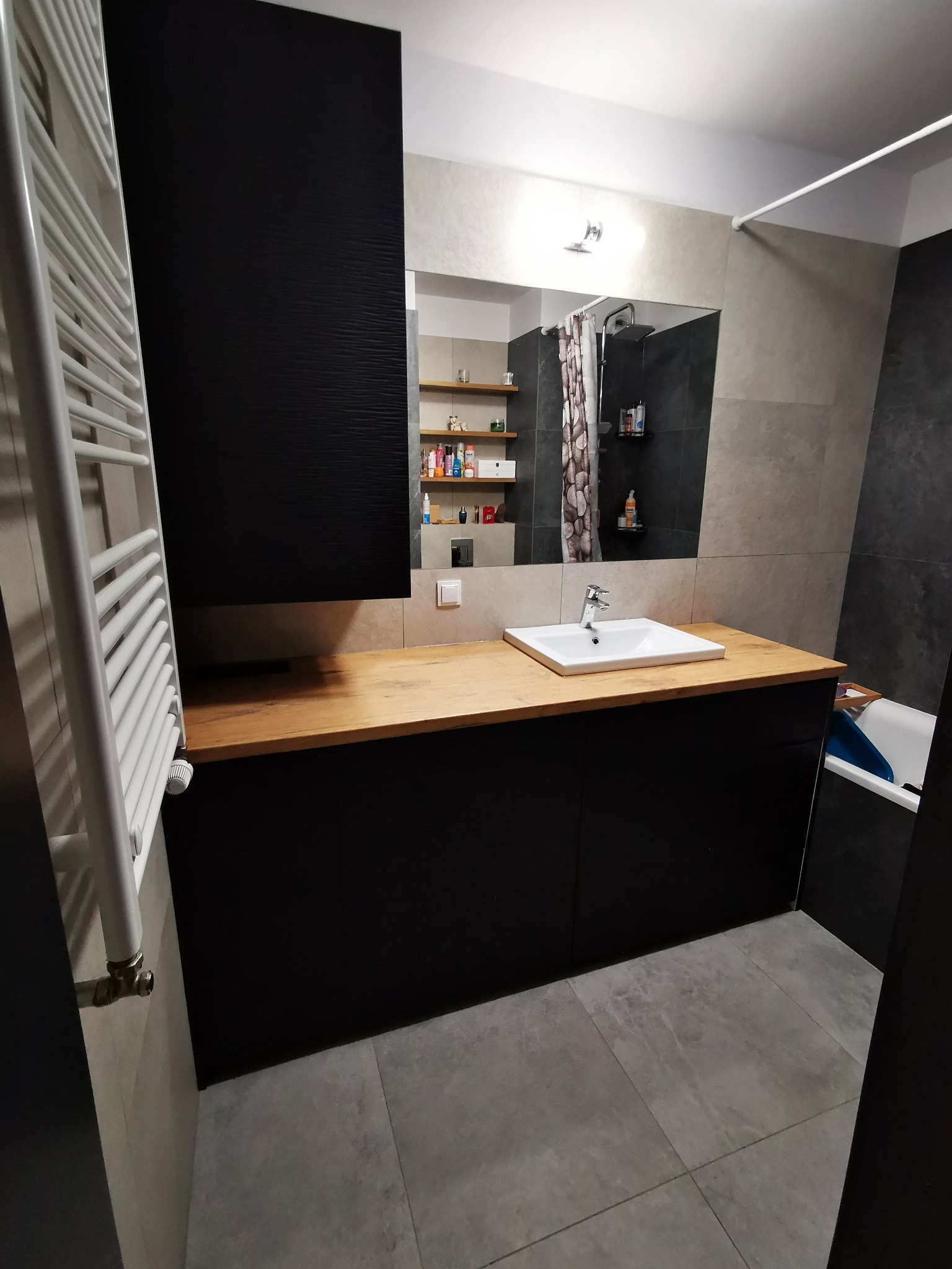 Nowoczesna łazienka z czarnymi szafkami, drewnianym blatem pod umywalką i grzejnikiem drabinkowym, w tle widoczna kabina prysznicowa.