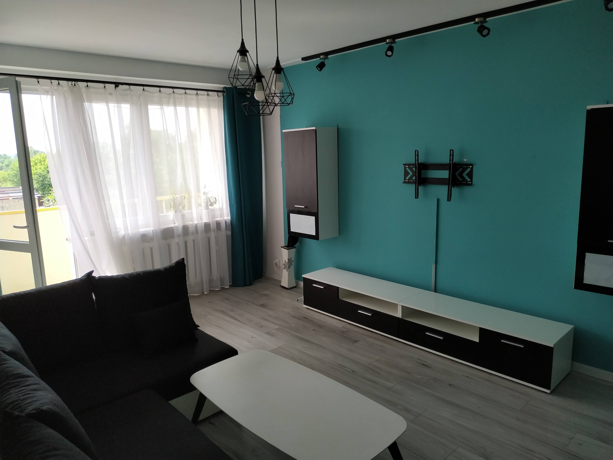 Turkusowa ściana w salonie z zamontowanym uchwytem TV, nowoczesne oświetlenie sufitowe i minimalistyczne meble w odcieniach bieli i czerni.