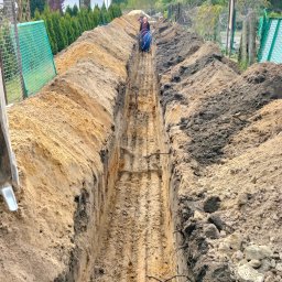 WJWBUD Usługi Sprzetowo Budowlane Jakub Wachowski - Długi, wąski wykop w ziemi z ułożonymi niebieskimi rurami w oddali, widoczne ślady po maszynie kopiącej na dnie wykopu, po bokach hałdy ziemi.