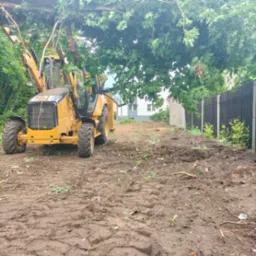 Żółta koparko-ładowarka CAT na placu budowy z częściowo wykarczowanym terenem, widoczne ślady opon w błocie, drzewa częściowo zasłaniają maszynę, w tle budynek mieszkalny i ogrodzenie.