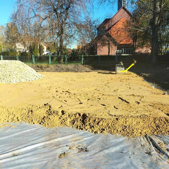 Przygotowany teren pod budowę z ubitym piaskiem, widoczna warstwa geowłókniny, kopiec żwiru i zagęszczarka wibracyjna.