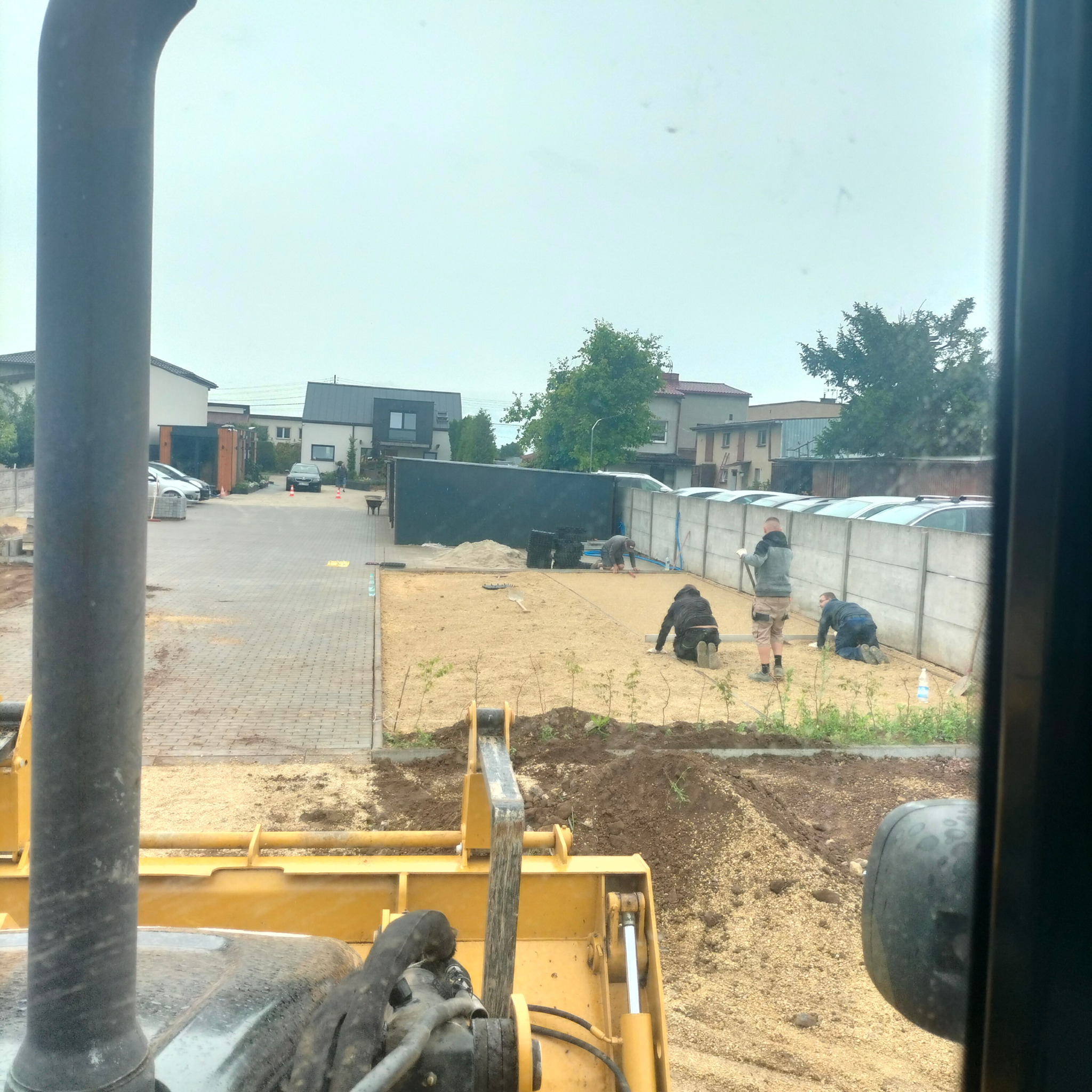 Widok z kabiny żółtej maszyny budowlanej na teren wyrównywany przez trzech pracowników, z widocznymi budynkami mieszkalnymi w tle.