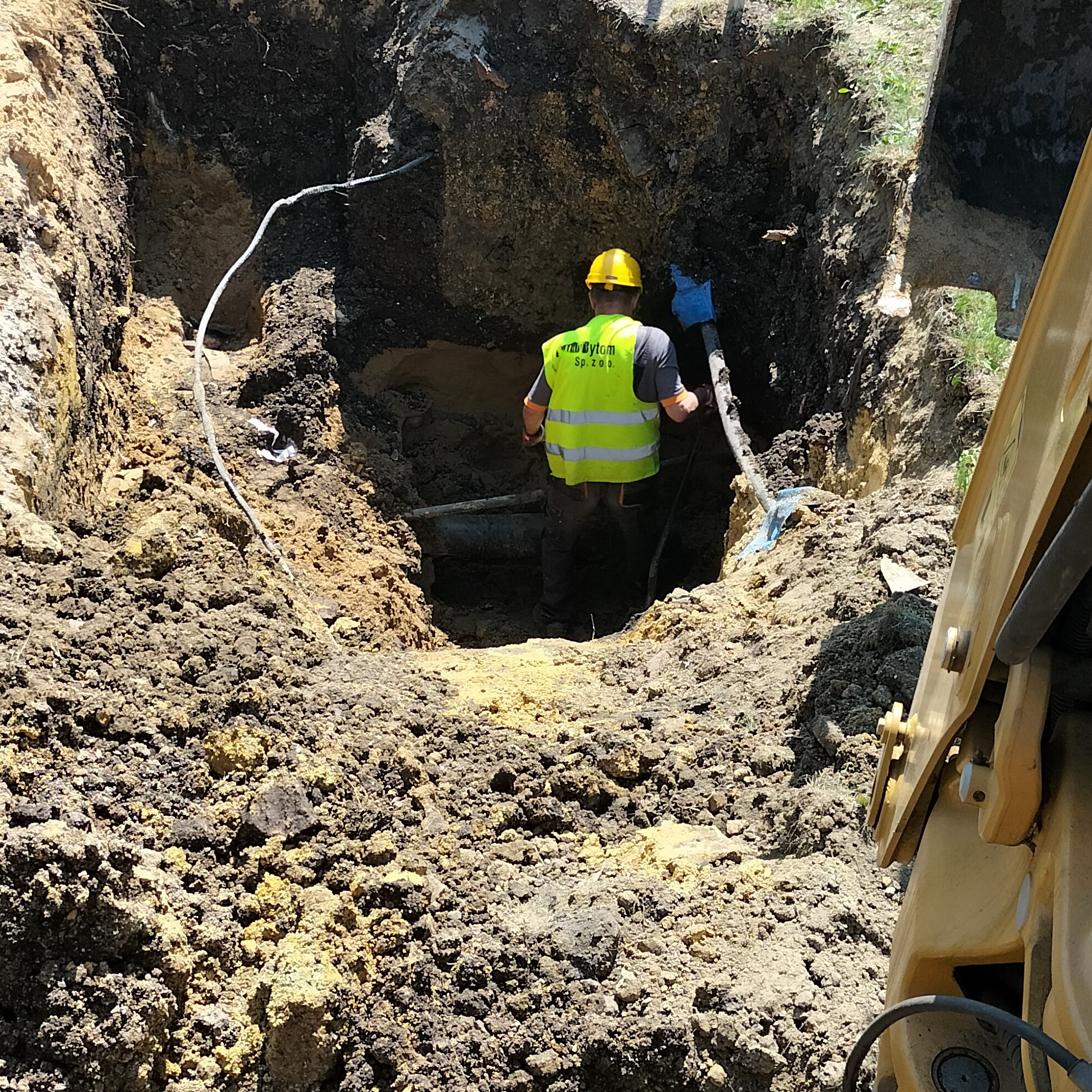 Pracownik w żółtym kasku i kamizelce odblaskowej stoi w głębokim wykopie, widoczne rury i kabel, fragment koparki z prawej strony.