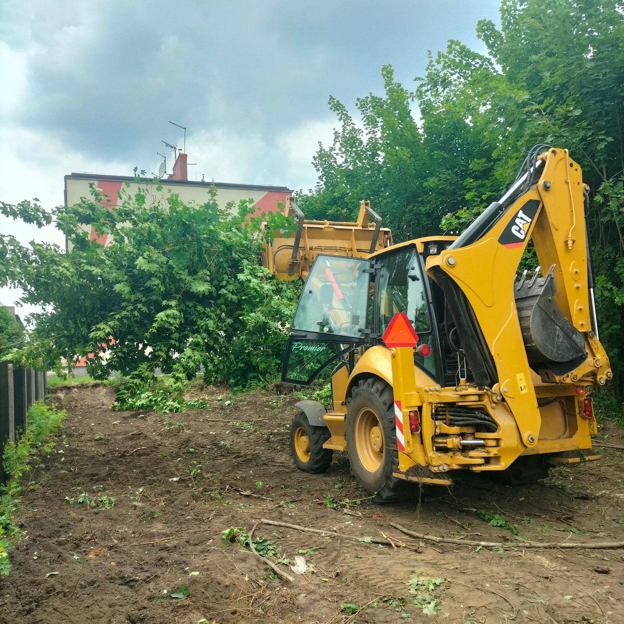 Żółta koparko-ładowarka CAT na uprzątniętym placu budowy, widoczne ślady opon w błotnistym gruncie, w tle budynek i drzewa pod pochmurnym niebem.