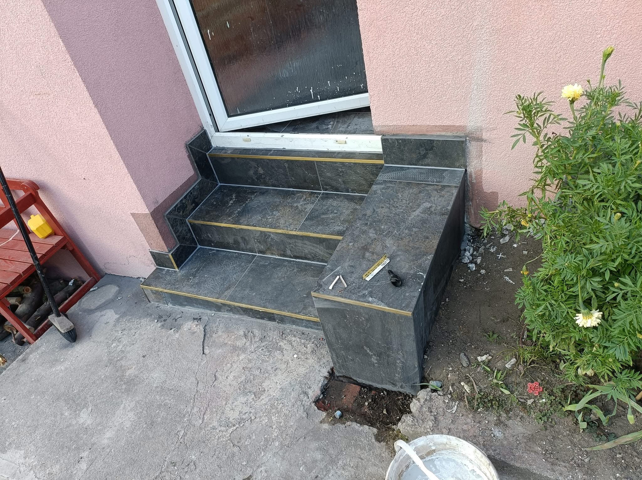 Wykończone schody wejściowe z szarych płytek z metalowymi listwami, widoczne narzędzia glazurnicze na górnym stopniu, obok roślina z żółtymi kwiatami, tło stanowi elewacja budynku w odcieniach różu.