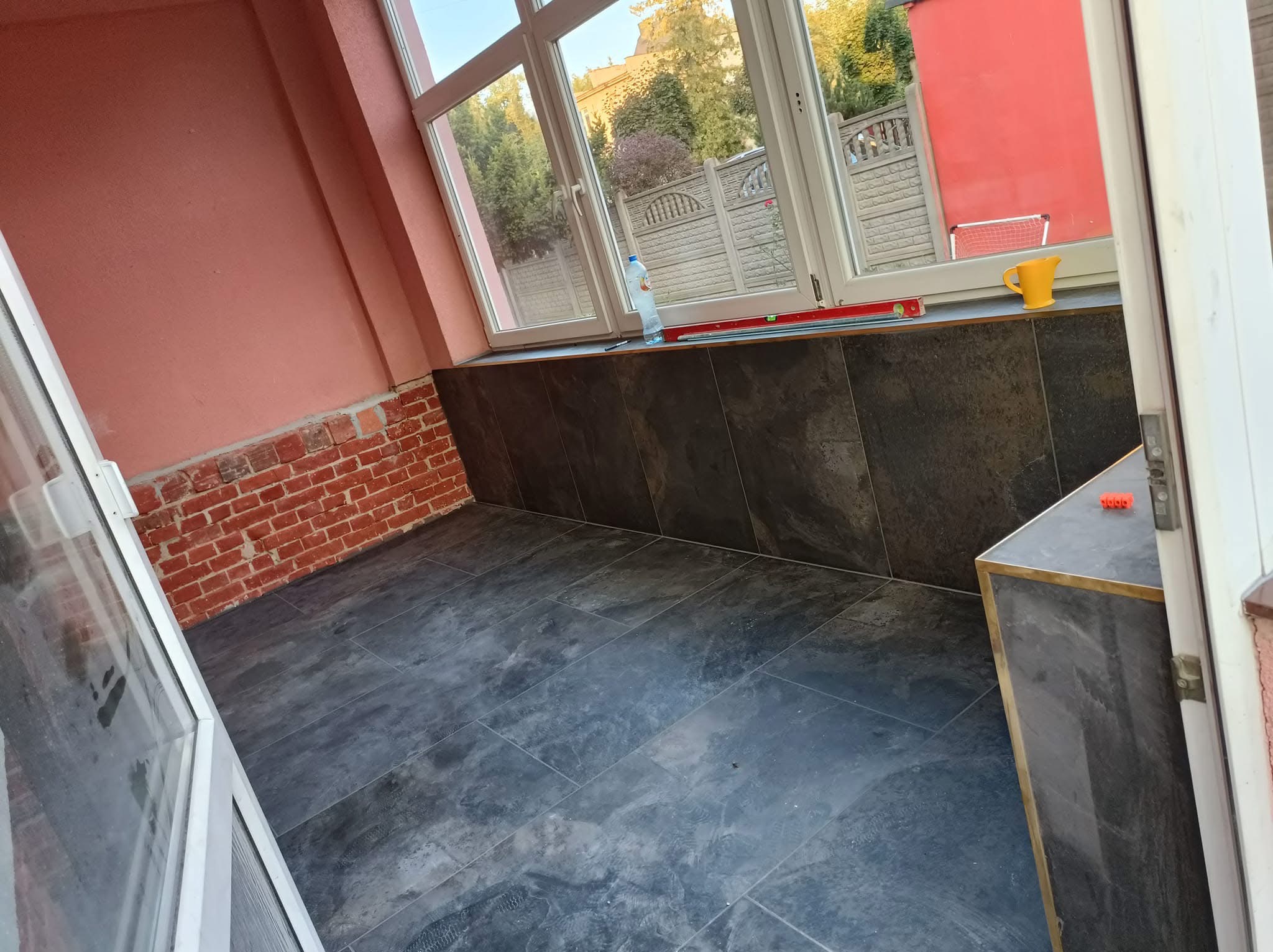 Wnętrze pomieszczenia w trakcie prac wykończeniowych: częściowo wyłożona ciemnymi płytkami ściana, surowa cegła na fragmencie ściany, widoczne płytki podłogowe, okno z widokiem na zewnątrz...