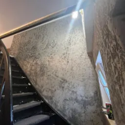 Ściana ze strukturą dekoracyjną w odcieniach szarości przy schodach. Widoczne okno z puszką napoju na parapecie. Czarna balustrada schodów.