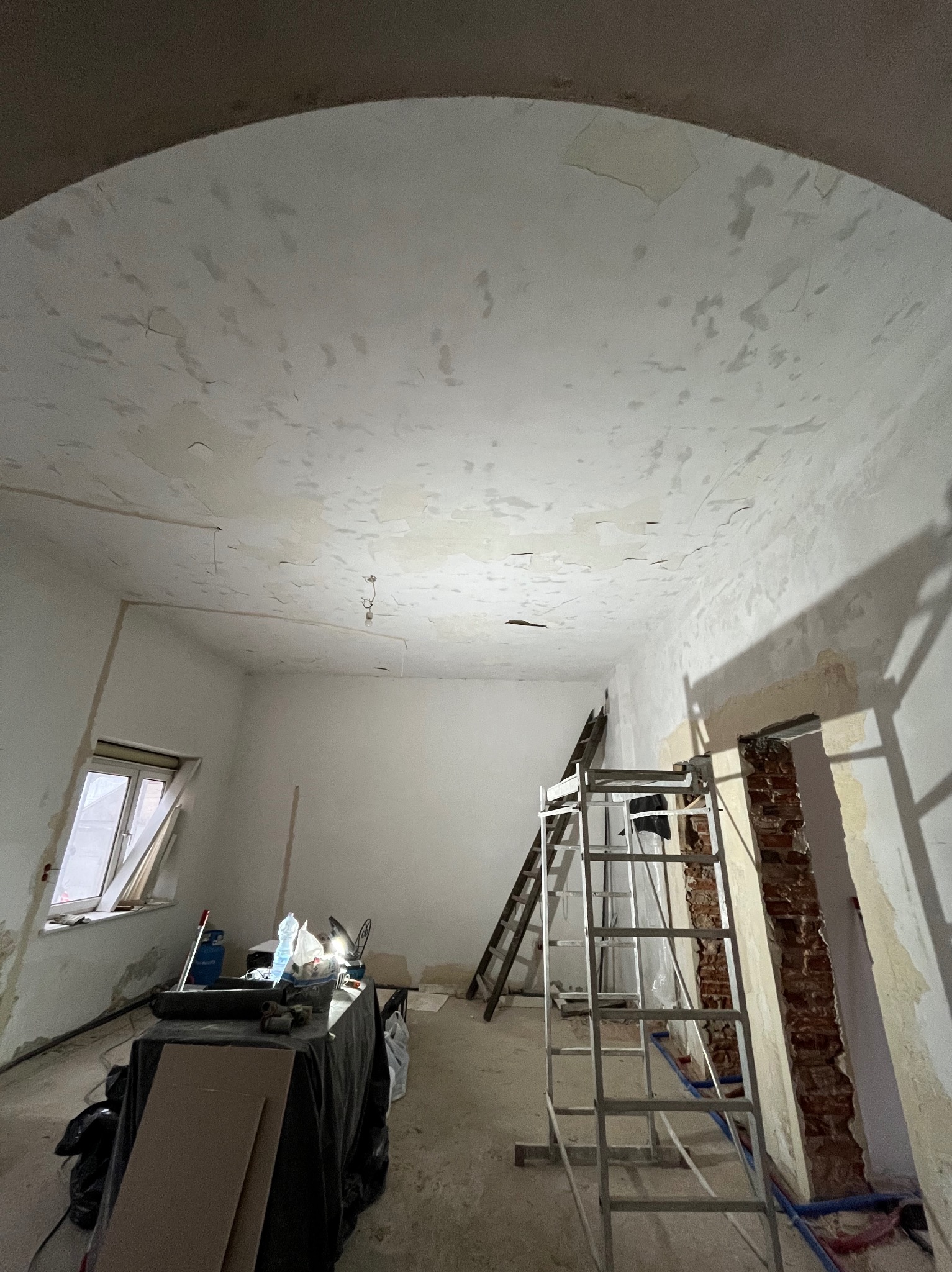 Pokój w trakcie remontu z widocznymi śladami po starych powłokach na suficie, dwa rodzaje drabin, stolik roboczy z narzędziami i materiałami budowlanymi, fragment odsłoniętej cegły w ścianie.
