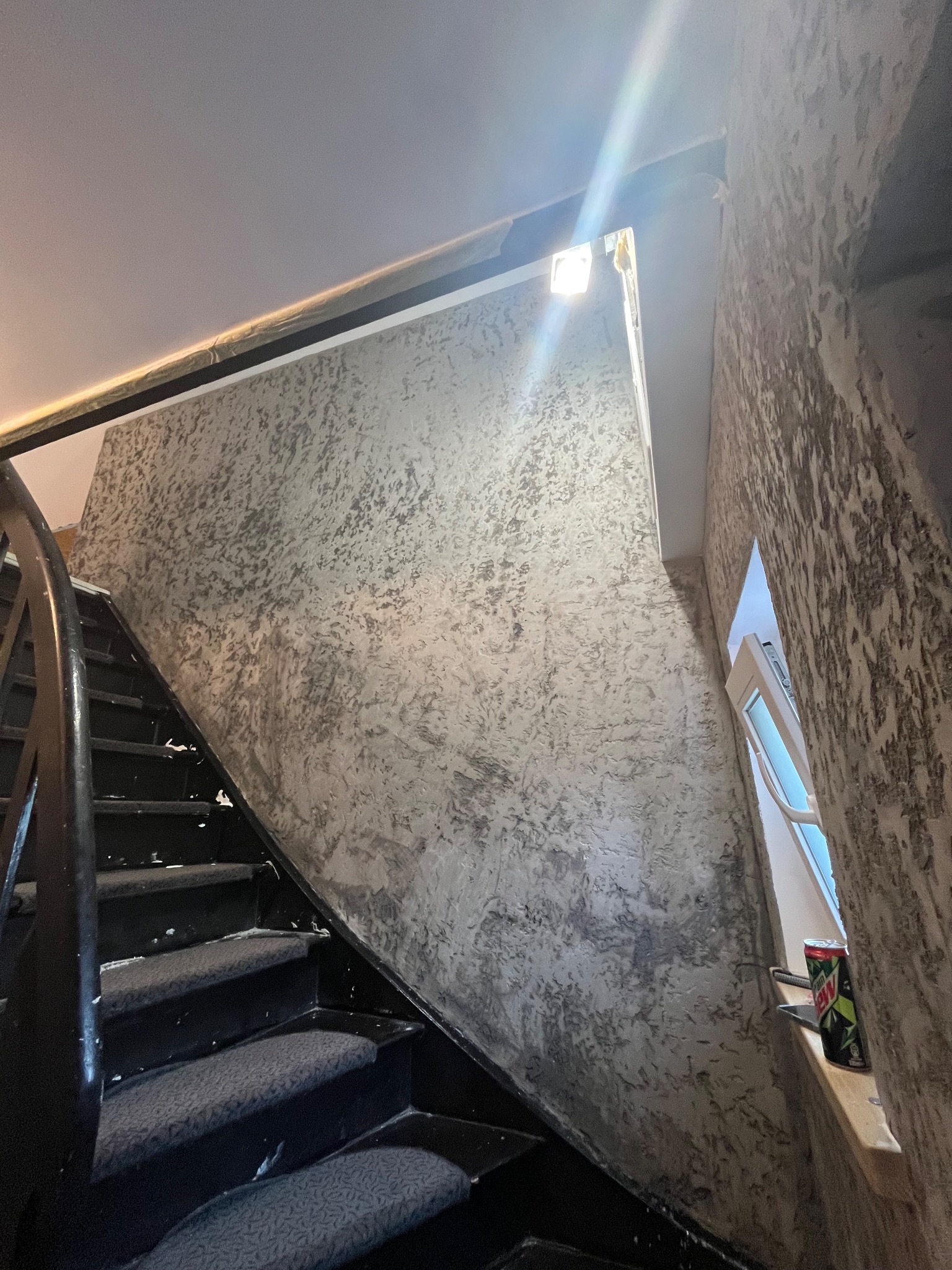 Ściana ze strukturą dekoracyjną w odcieniach szarości przy schodach. Widoczne okno z puszką napoju na parapecie. Czarna balustrada schodów.