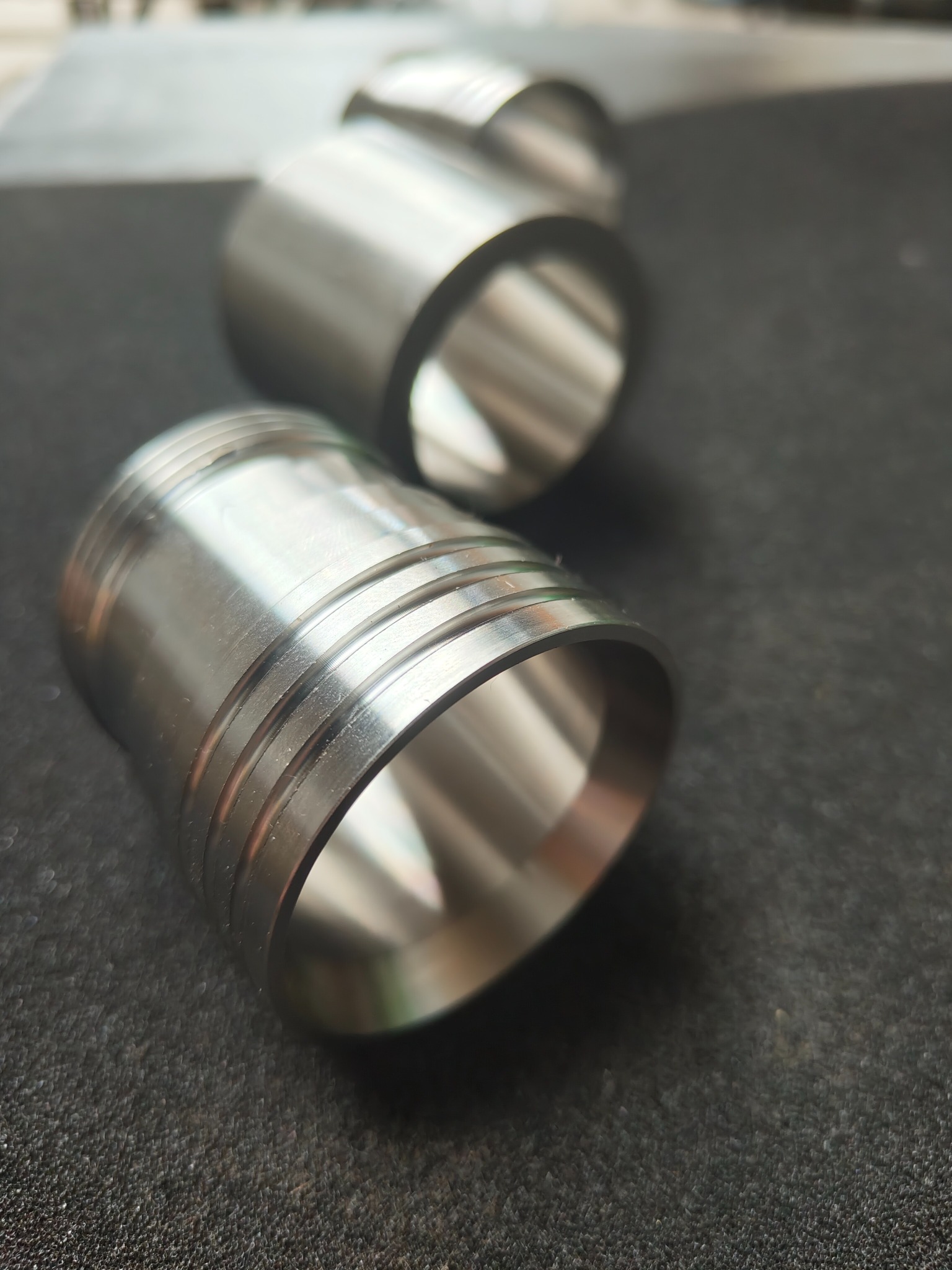 Precyzyjnie wykonane, metalowe tuleje z rowkami, leżące na ciemnoszarym, teksturowanym podłożu, ukazujące detale obróbki.