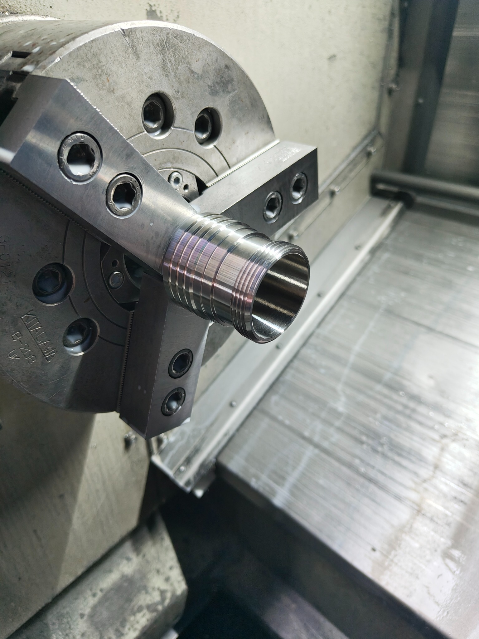 Metalowy element rurowy z gwintem, obrabiany w uchwycie tokarskim CNC, widoczne ślady obróbki i opalizujące zabarwienie metalu.