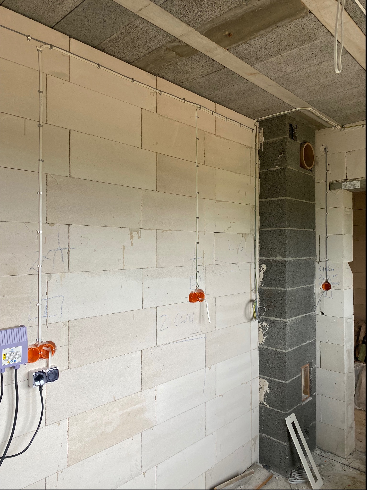 Wnętrze w trakcie prac elektrycznych: widoczne kable w peszlach mocowane do ściany z betonu komórkowego, puszki instalacyjne w kolorze pomarańczowym, gniazdka oraz szara kolumna kominowa z otworem...