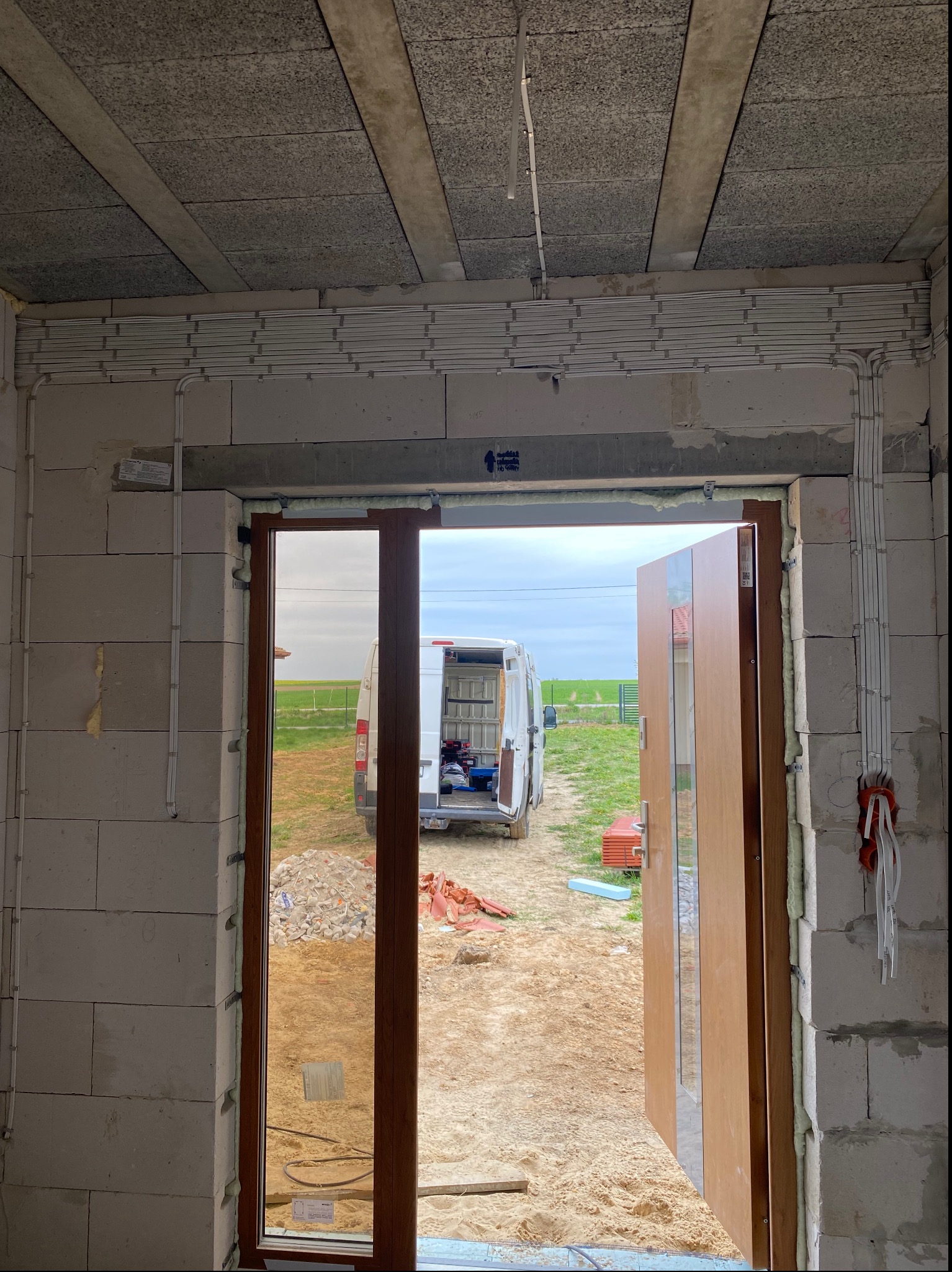 Wnętrze surowego budynku w trakcie prac wykończeniowych: widoczne kable elektryczne ułożone w korytach na ścianie z betonu komórkowego, otwarte drzwi wejściowe z widokiem na podwórko z zaparkowanym...