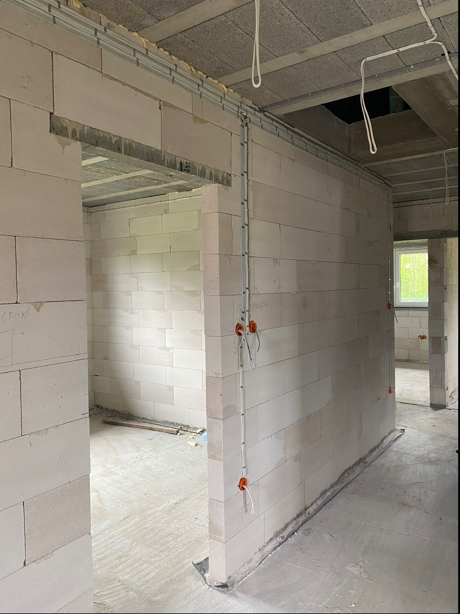 Surowy stan wnętrza z widocznymi przewodami elektrycznymi w peszlach, zamontowanymi puszkami instalacyjnymi, oraz szynami montażowymi pod sufitem w nowym budynku z bloczków.