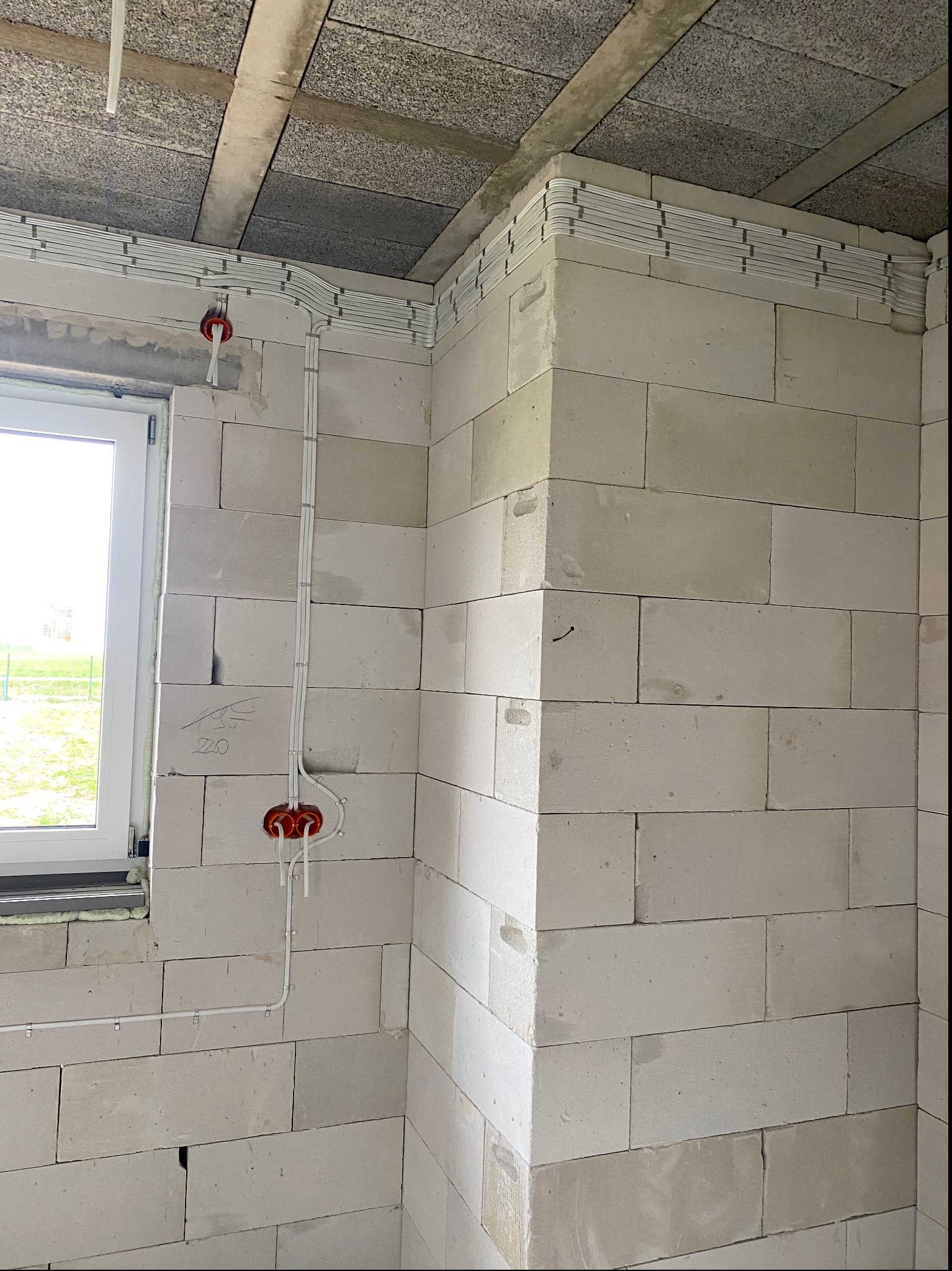 Wewnętrzna instalacja elektryczna w surowym budynku z białych bloczków, widoczne kable prowadzone w listwach na ścianach i pod sufitem, puszki elektryczne w kolorze pomarańczowym, okno z widokiem...