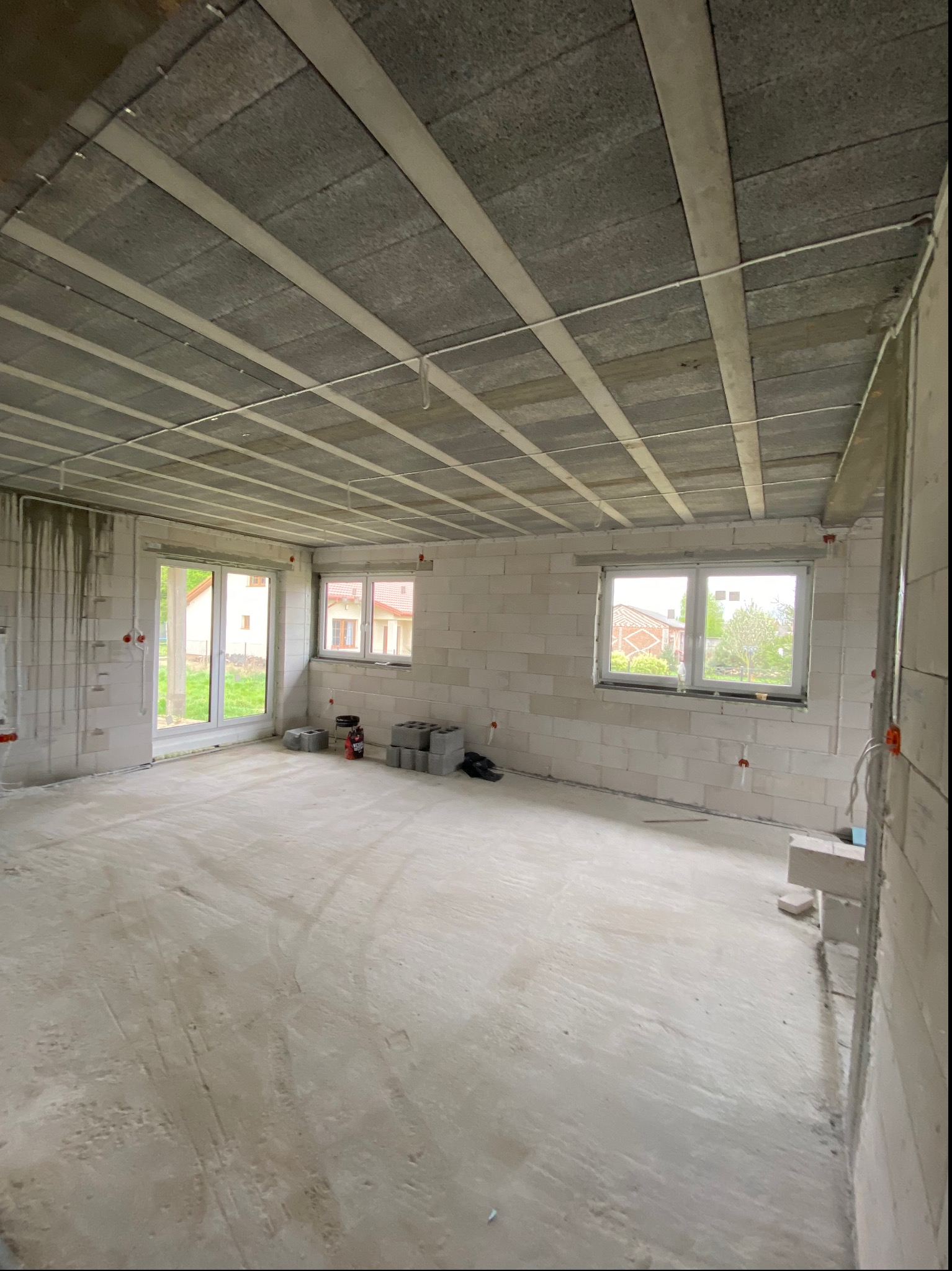 Surowe wnętrze domu w trakcie budowy z widocznymi przewodami elektrycznymi wystającymi ze ścian i sufitu, pustaki na podłodze i oknami na zewnątrz.