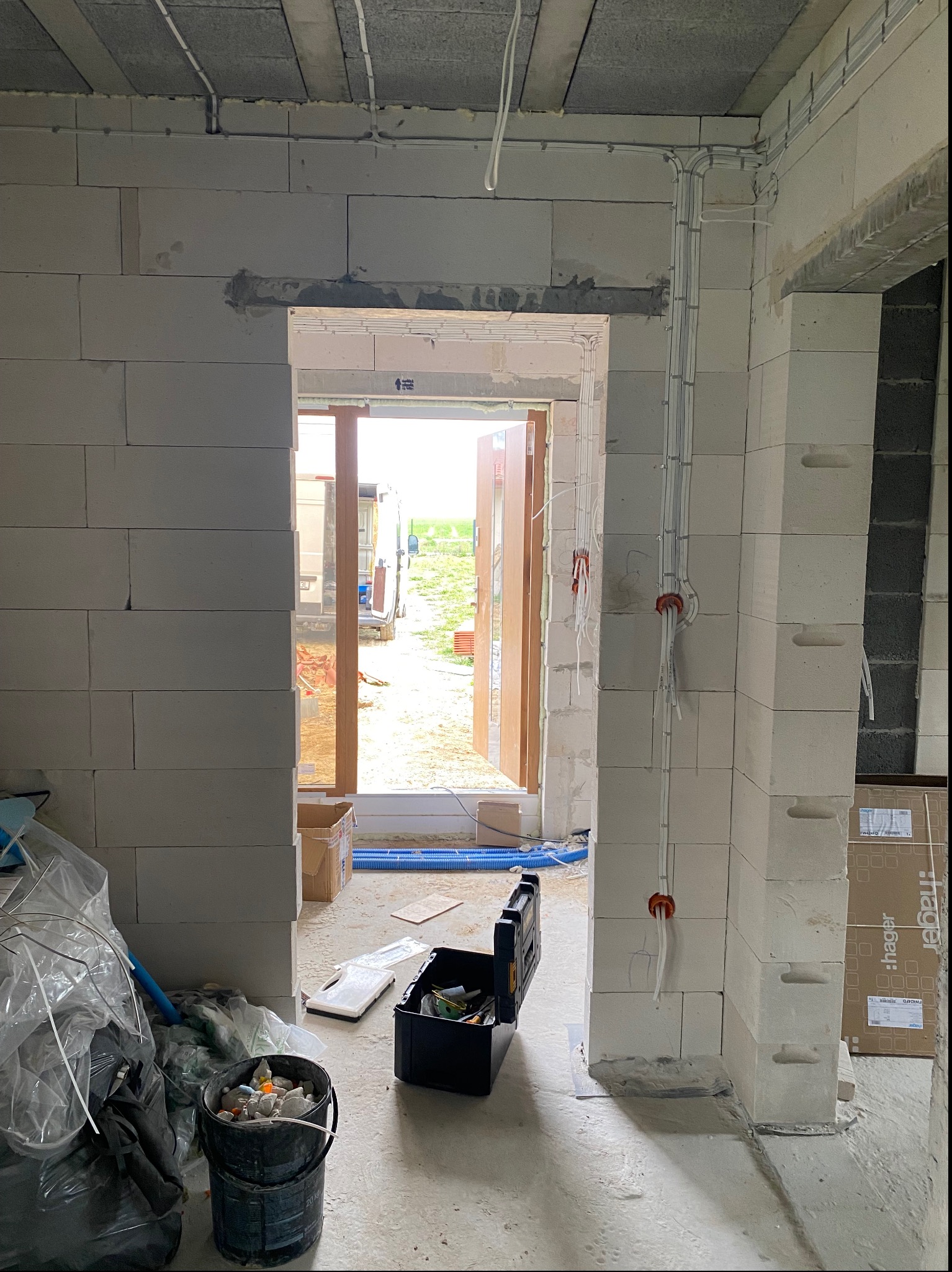 Wnętrze surowego budynku w trakcie prac instalacyjnych: widoczne przewody elektryczne prowadzone po ścianach z pustaków, skrzynka narzędziowa i wiadro z gruzem na podłodze, otwarte drzwi z widokiem...