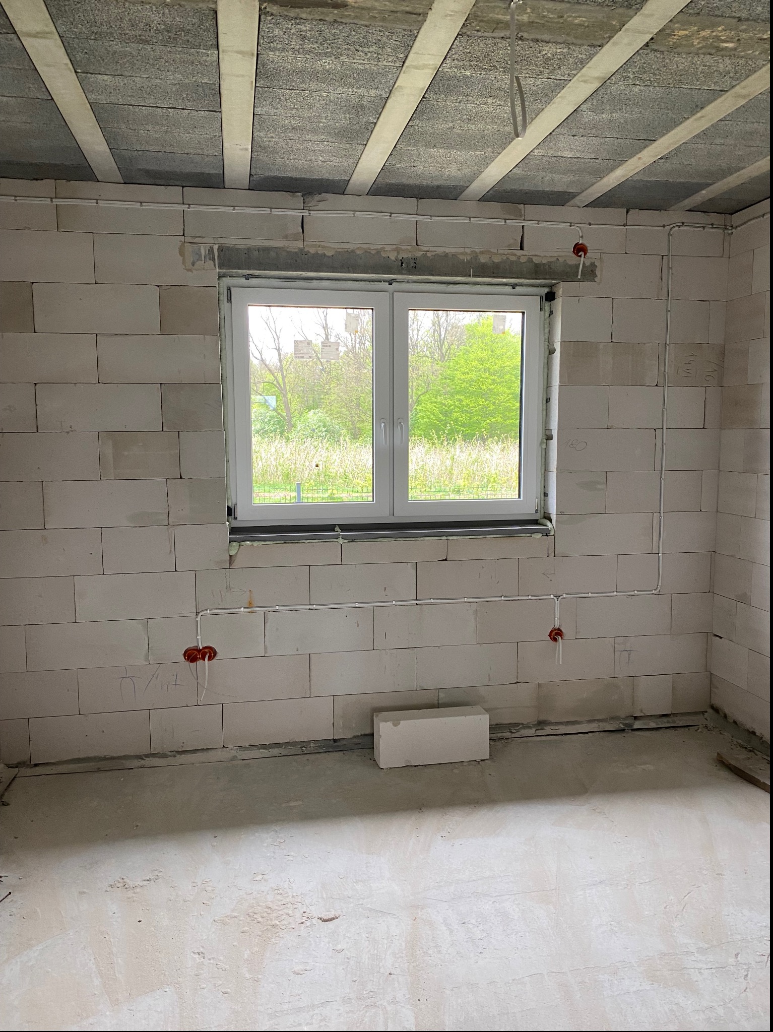 Surowy stan wnętrza z widoczną instalacją elektryczną w ścianach z betonu komórkowego, okno z widokiem na zieleń, sufit z elementami konstrukcyjnymi.