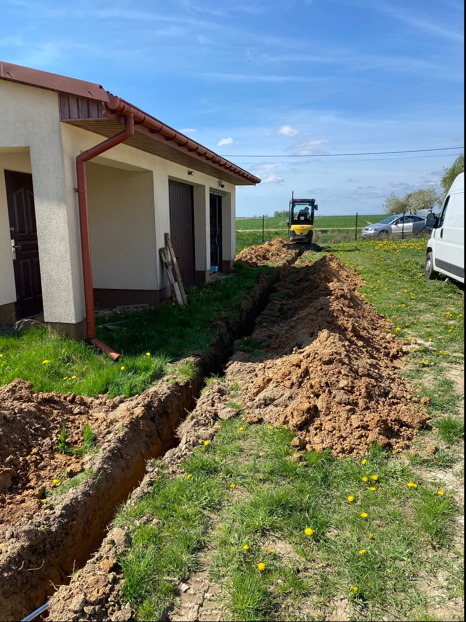 Wykopany rów na trawniku przed budynkiem, z żółtym mini-koparką w tle i stertą ziemi po bokach, sugerujący prace ziemne związane z instalacją.