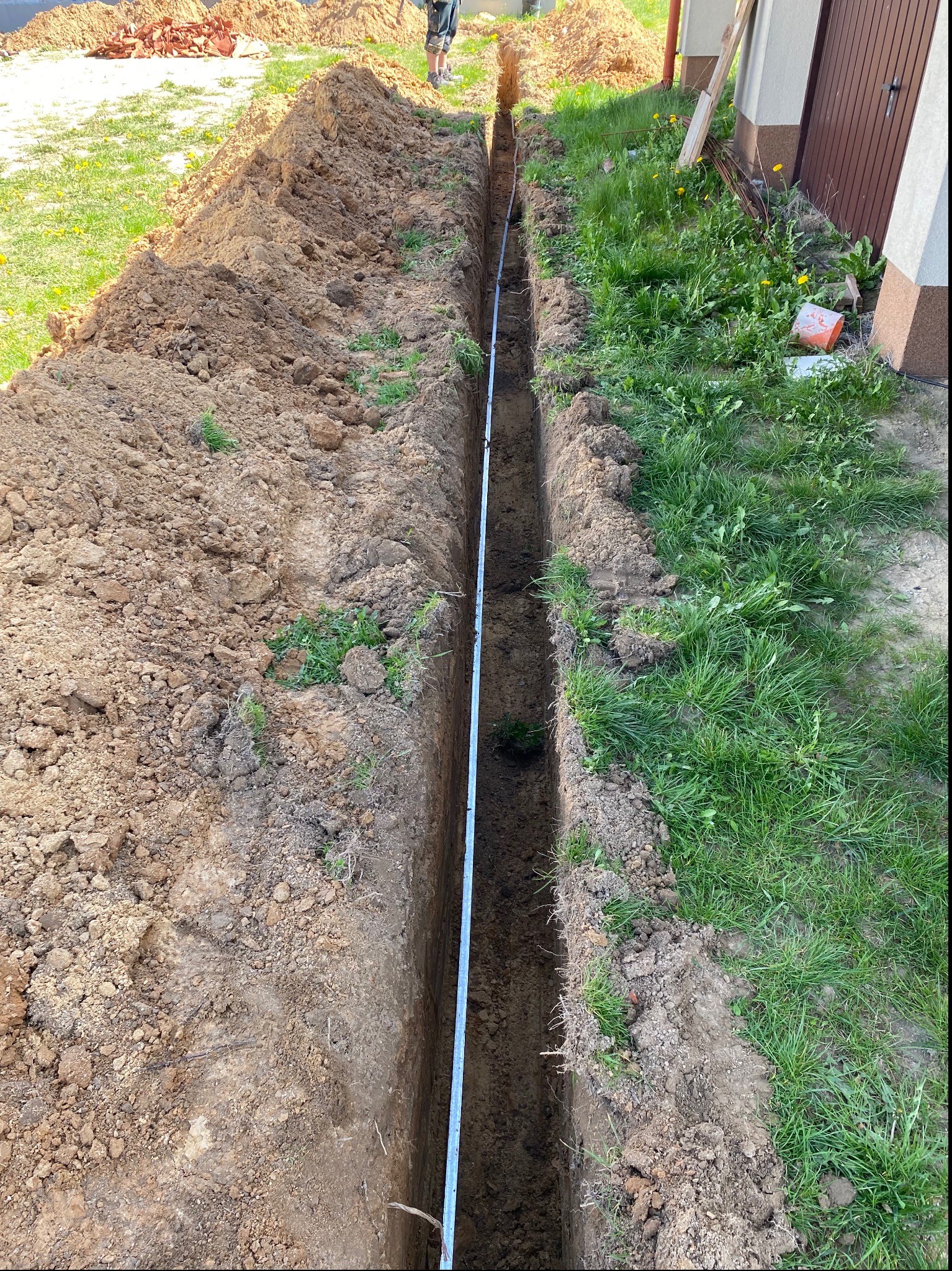 Wykopany wąski rów w ziemi, prawdopodobnie pod kabel elektryczny, z widoczną miarką wewnątrz rowu i stertą ziemi po obu stronach.