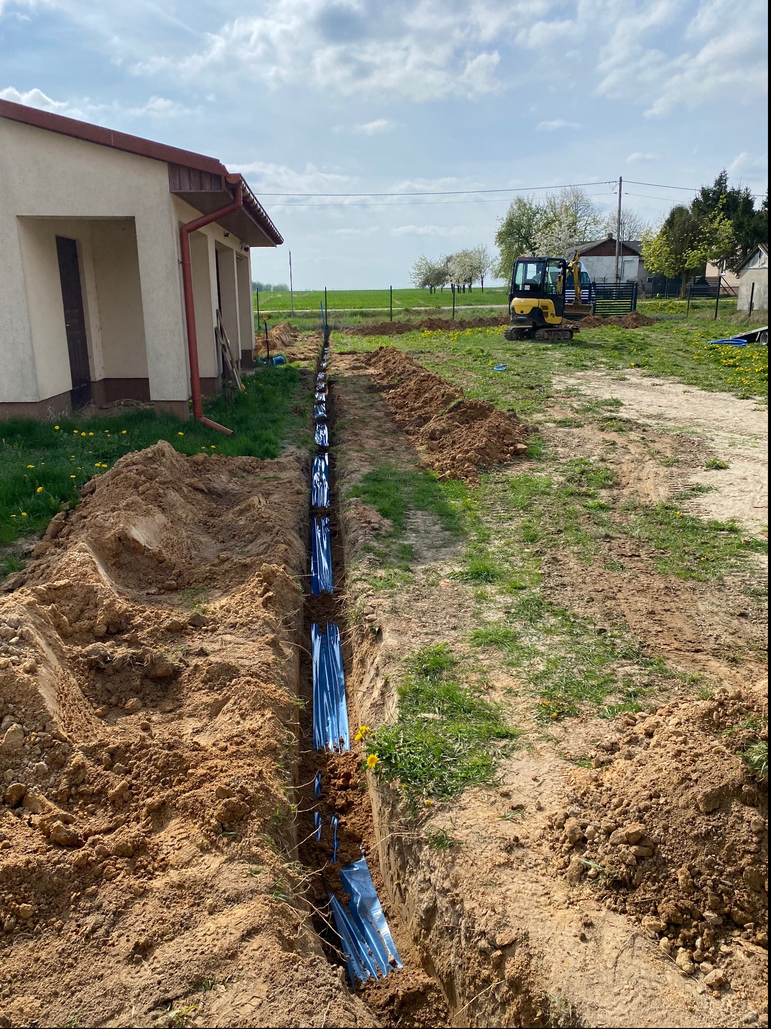 Wkop pod ziemią z niebieskimi rurami ochronnymi, wykopany wzdłuż budynku, z koparką w tle na trawniku.