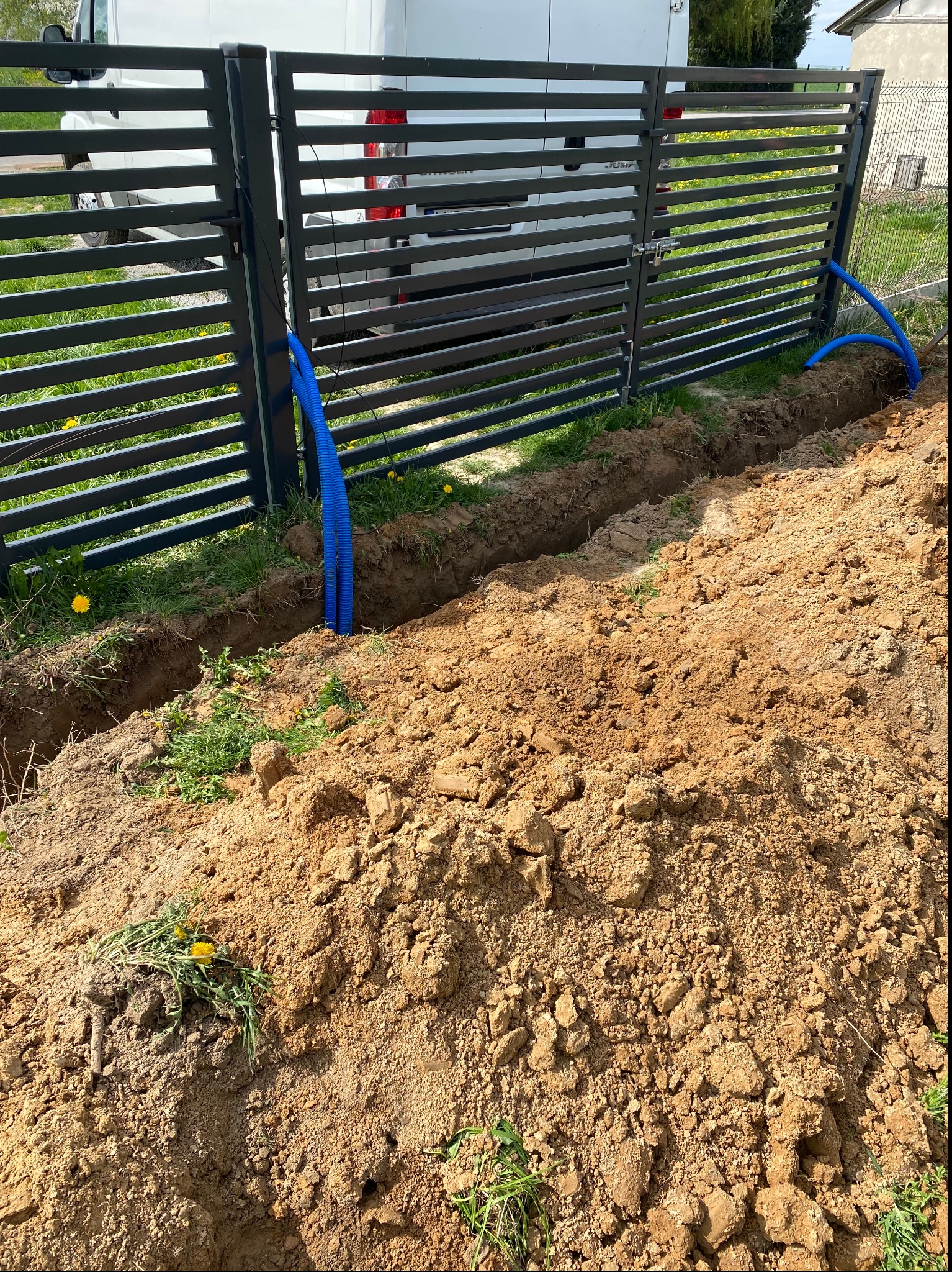 Niebieskie rury ochronne ułożone w wykopie ziemnym wzdłuż metalowego ogrodzenia posesji. Na tle zaparkowany biały bus.