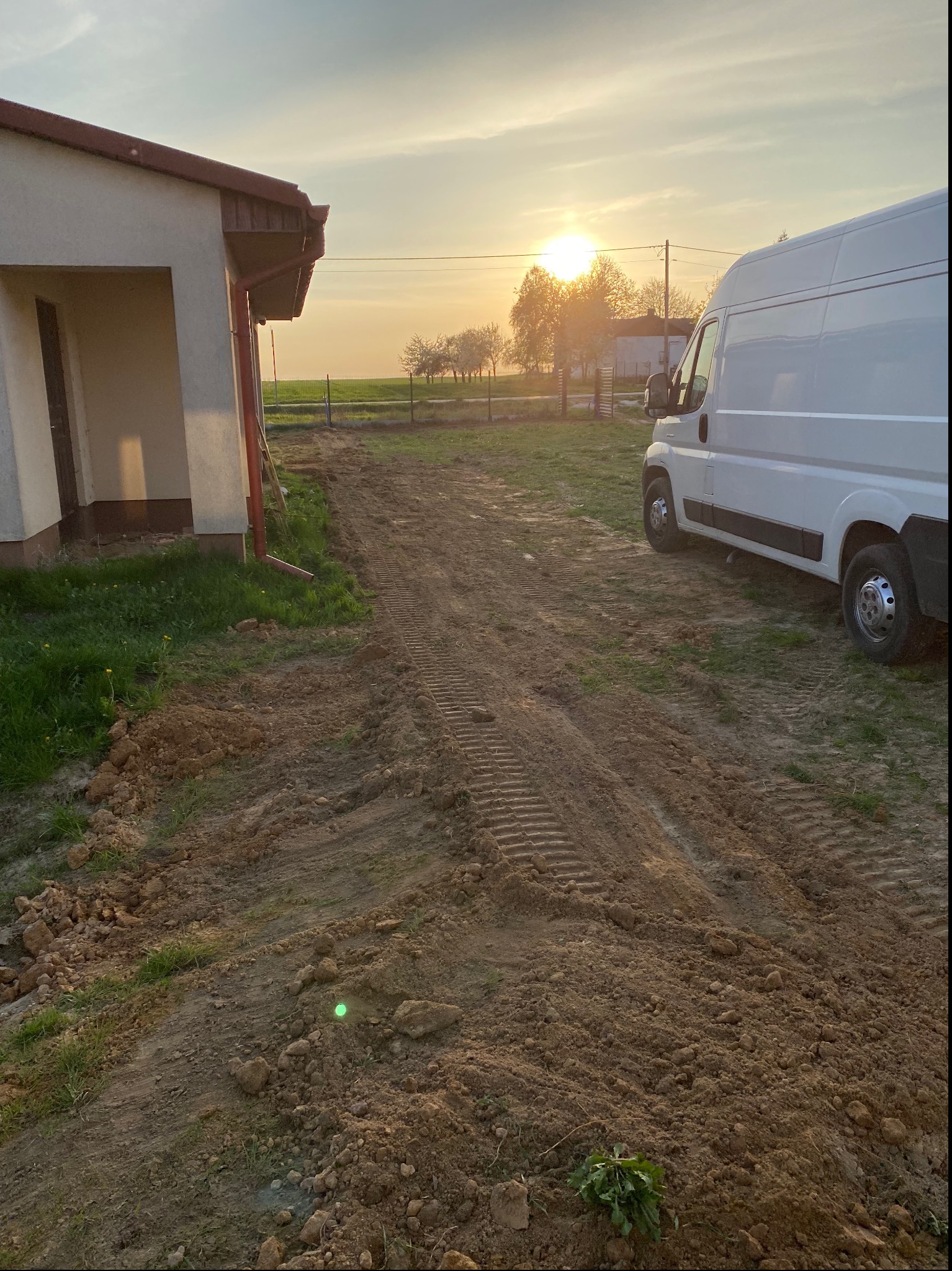 Przygotowanie terenu pod instalację: wyrównana ziemia z śladami opon, biały samochód dostawczy w tle, budynek z rynnami i zachodzące słońce.