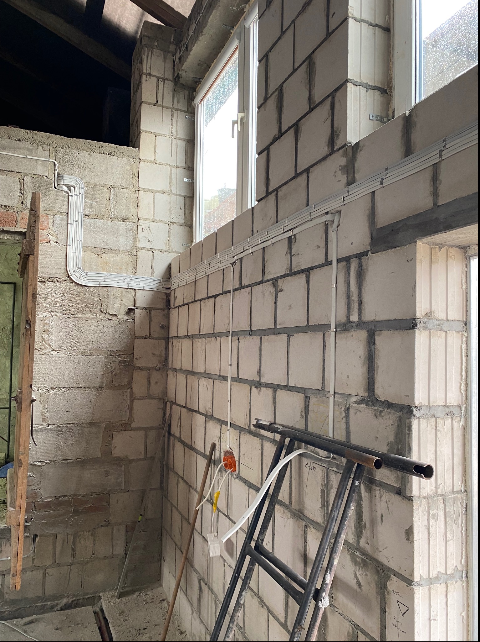 Wewnętrzne okablowanie elektryczne w białych listwach na surowych ścianach z betonu komórkowego w trakcie budowy, widoczna puszka instalacyjna i drabina.