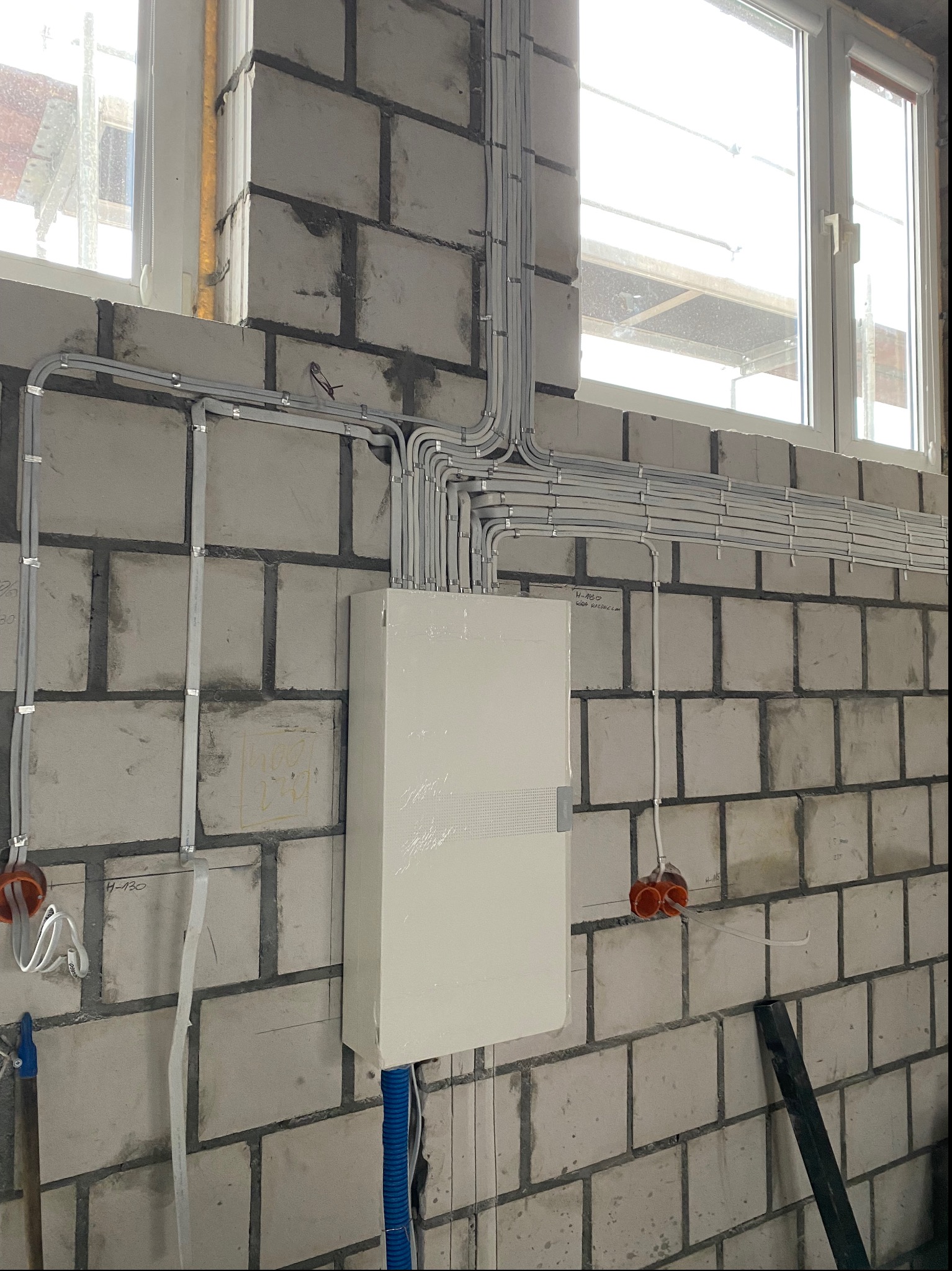 Rozdzielnica elektryczna z licznymi przewodami ułożonymi w peszlach na surowej ścianie z betonu komórkowego, widoczne okno i fragment rusztowania na zewnątrz.