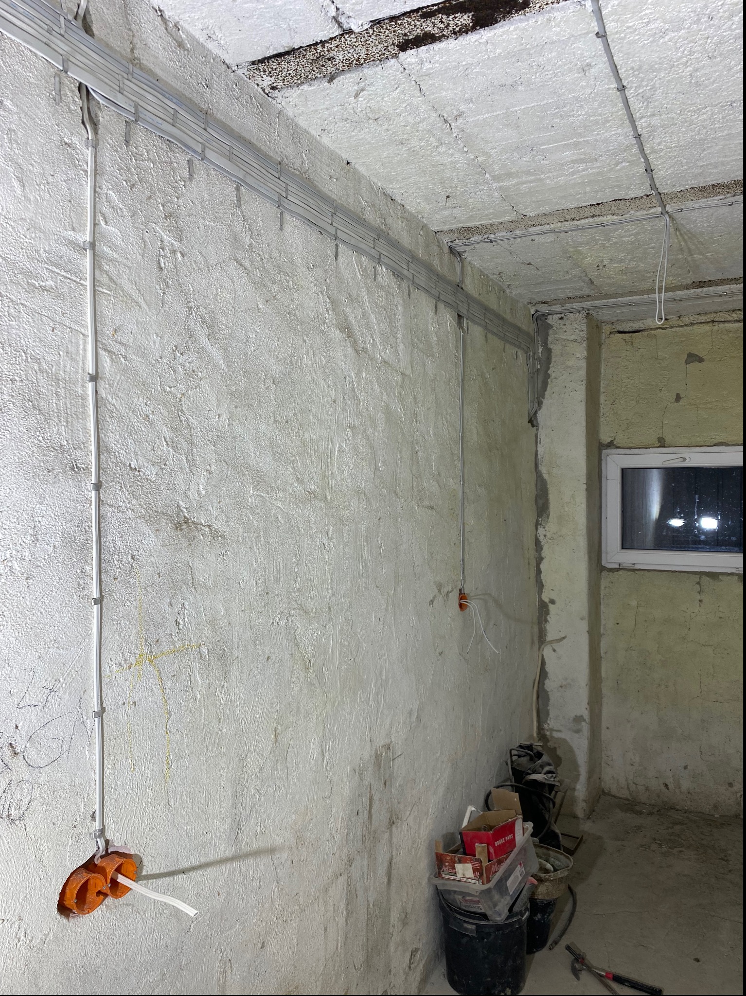 Wnętrze w Wojciechowie w trakcie remontu: widoczne kable elektryczne prowadzone w korytkach na ścianie i suficie, puszki instalacyjne w ścianie, surowy tynk i okno w tle.