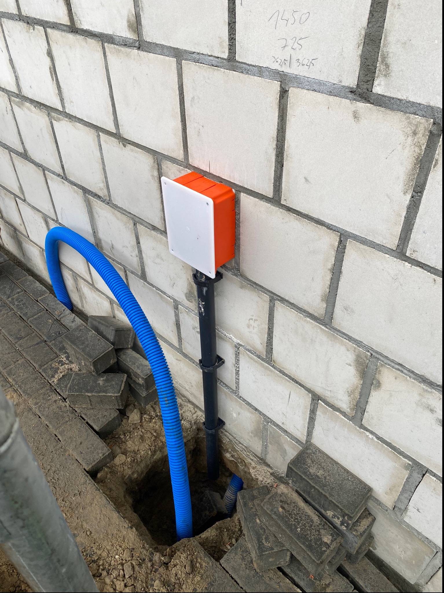 Pomarańczowa skrzynka elektryczna z białą pokrywą zamocowana na ścianie z betonu komórkowego, z niebieską rurą karbowaną prowadzącą do wykopu w ziemi, obok ułożona kostka brukowa.
