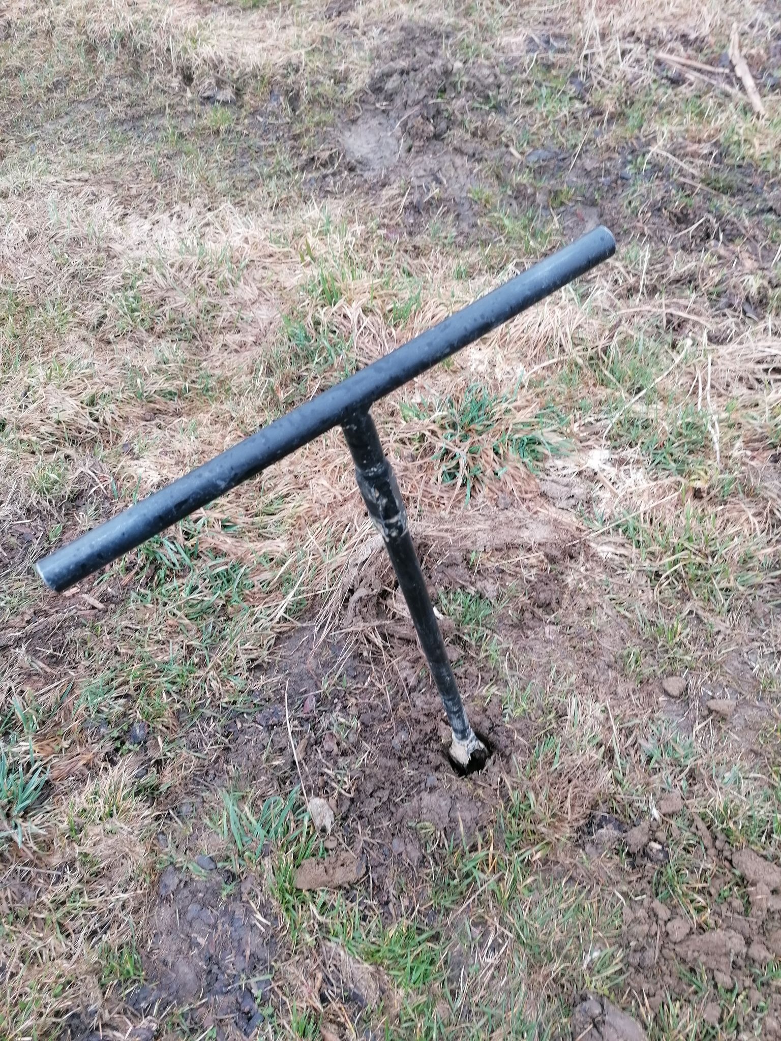 Wiertło glebowe w ziemi, widoczny otwór po pobraniu próbki, trawa i błoto w tle.