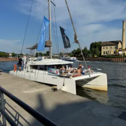 Biały katamaran 'Felicity' zacumowany przy nabrzeżu, z grupą pasażerów na pokładzie, widoczne flagi firmowe i budynek z kominem w tle.