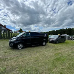 Dwa busy, czarny i szary, stoją na trawiastym terenie pod błękitnym niebem z chmurami; czarny bus z widocznym logo firmy na boku.