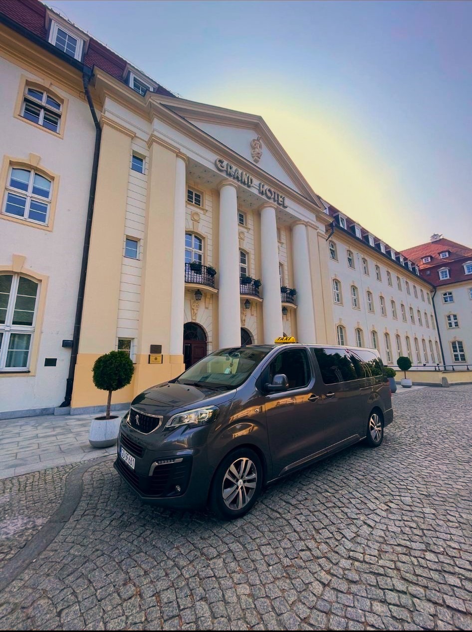 Szary bus z napisem TAXI na dachu zaparkowany na brukowanej drodze przed hotelem Grand Hotel z kolumnami.