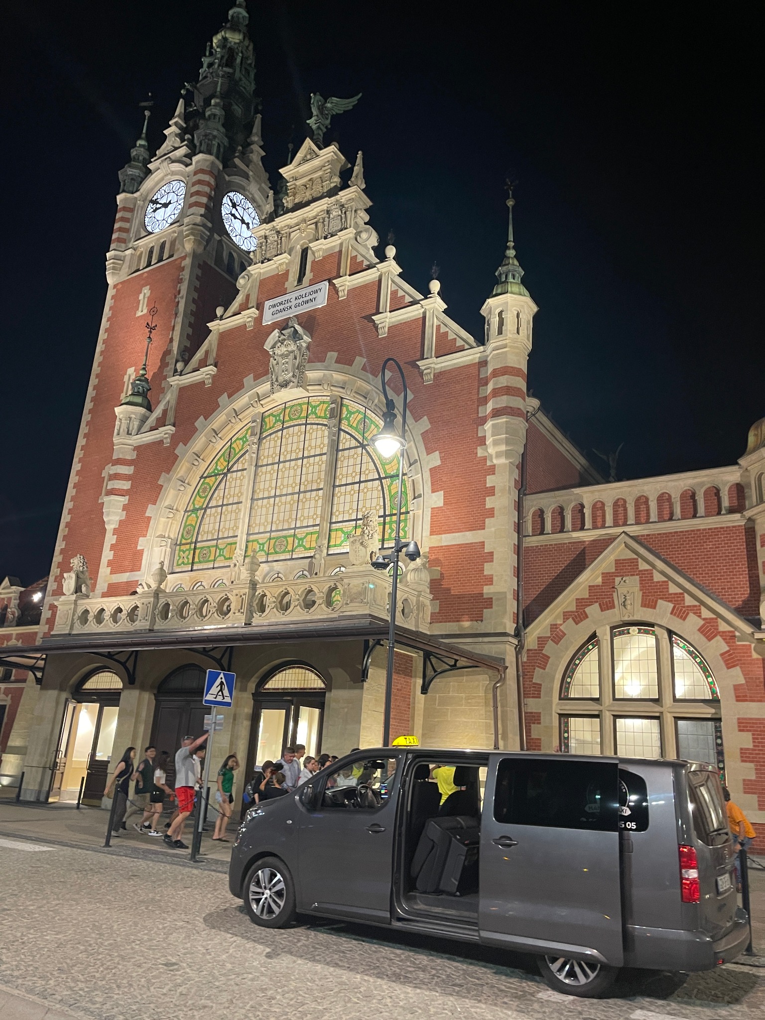 Szary bus z otwartymi drzwiami na tle budynku dworca kolejowego Gdańsk Główny nocą, pasażerowie w pobliżu.