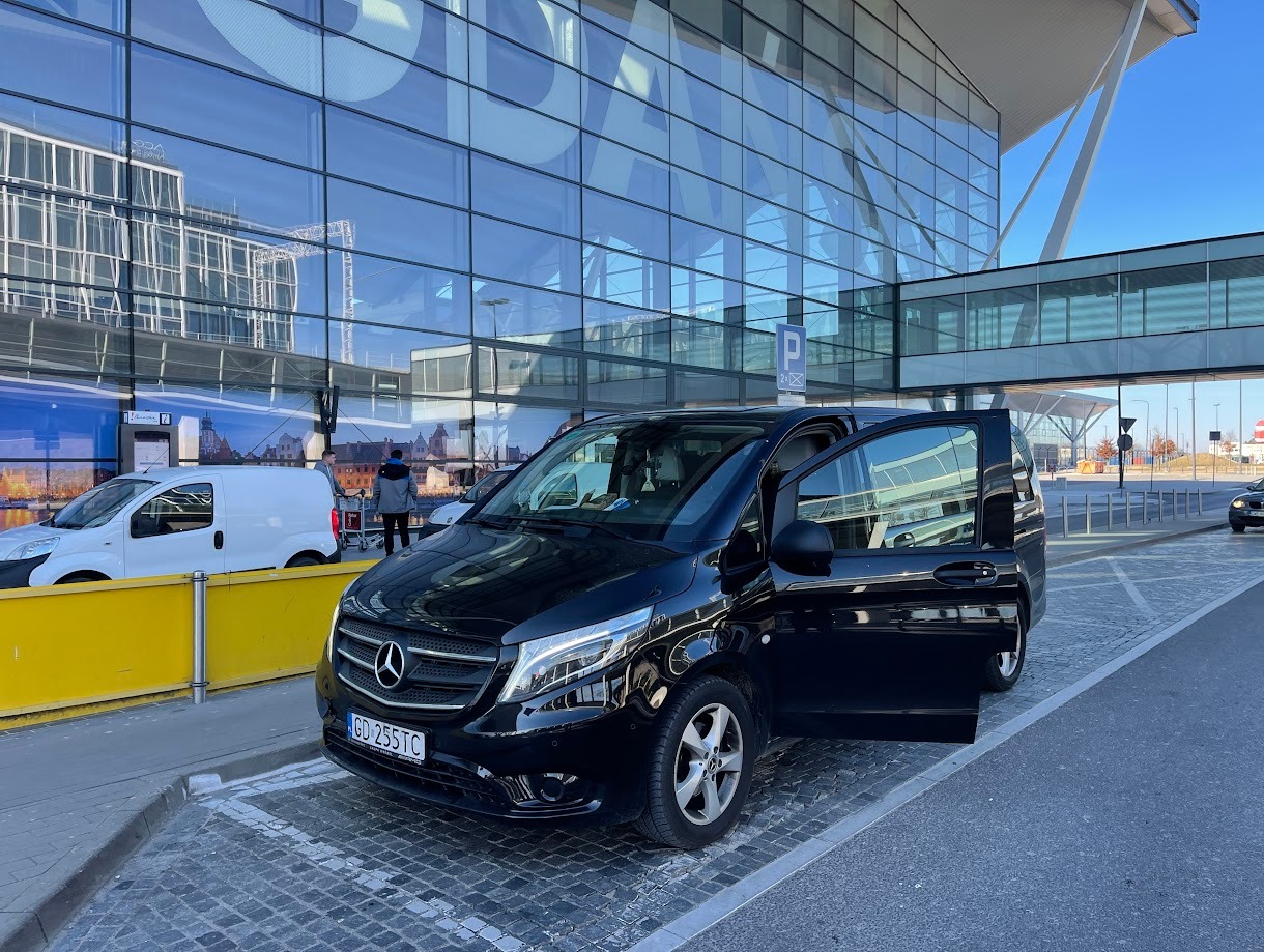 Czarny Mercedes Vito z otwartymi drzwiami na tle nowoczesnego budynku lotniska z szklaną fasadą i znakiem parkingu.