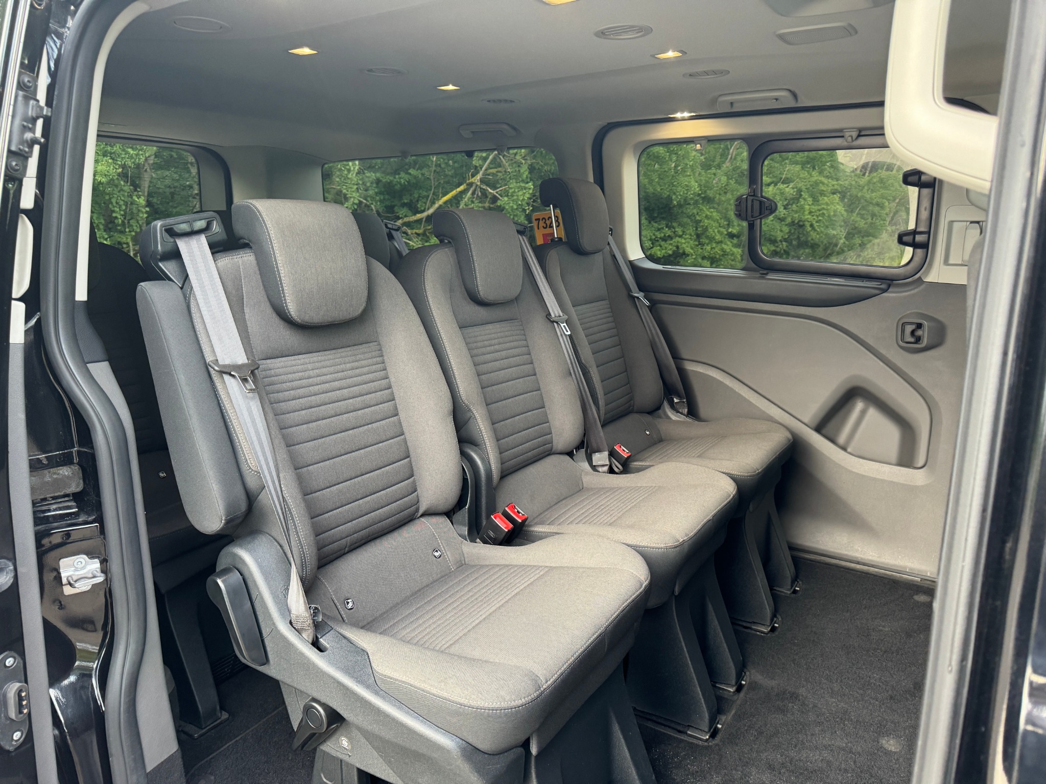 Wnętrze busa pasażerskiego z trzema rzędami szarych foteli z pasami bezpieczeństwa, widok z otwartych drzwi na zewnątrz, gdzie widać zieleń drzew.