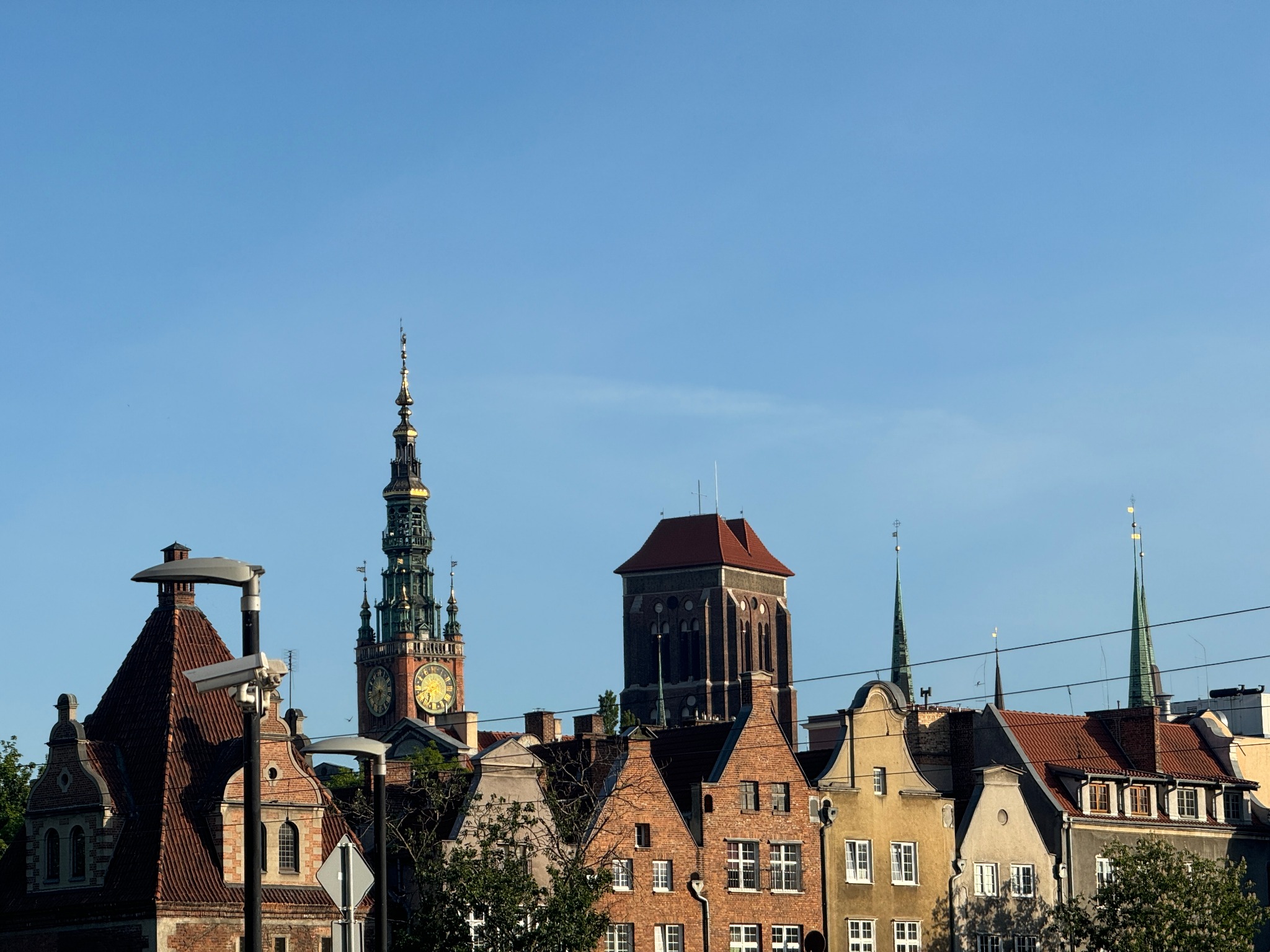 Panorama Gdańska z widokiem na zabytkowe kamienice i wieżę zegarową kościoła Mariackiego, sfotografowana w słoneczny dzień z perspektywy ulicy, z widocznymi przewodami elektrycznymi na pierwszym...