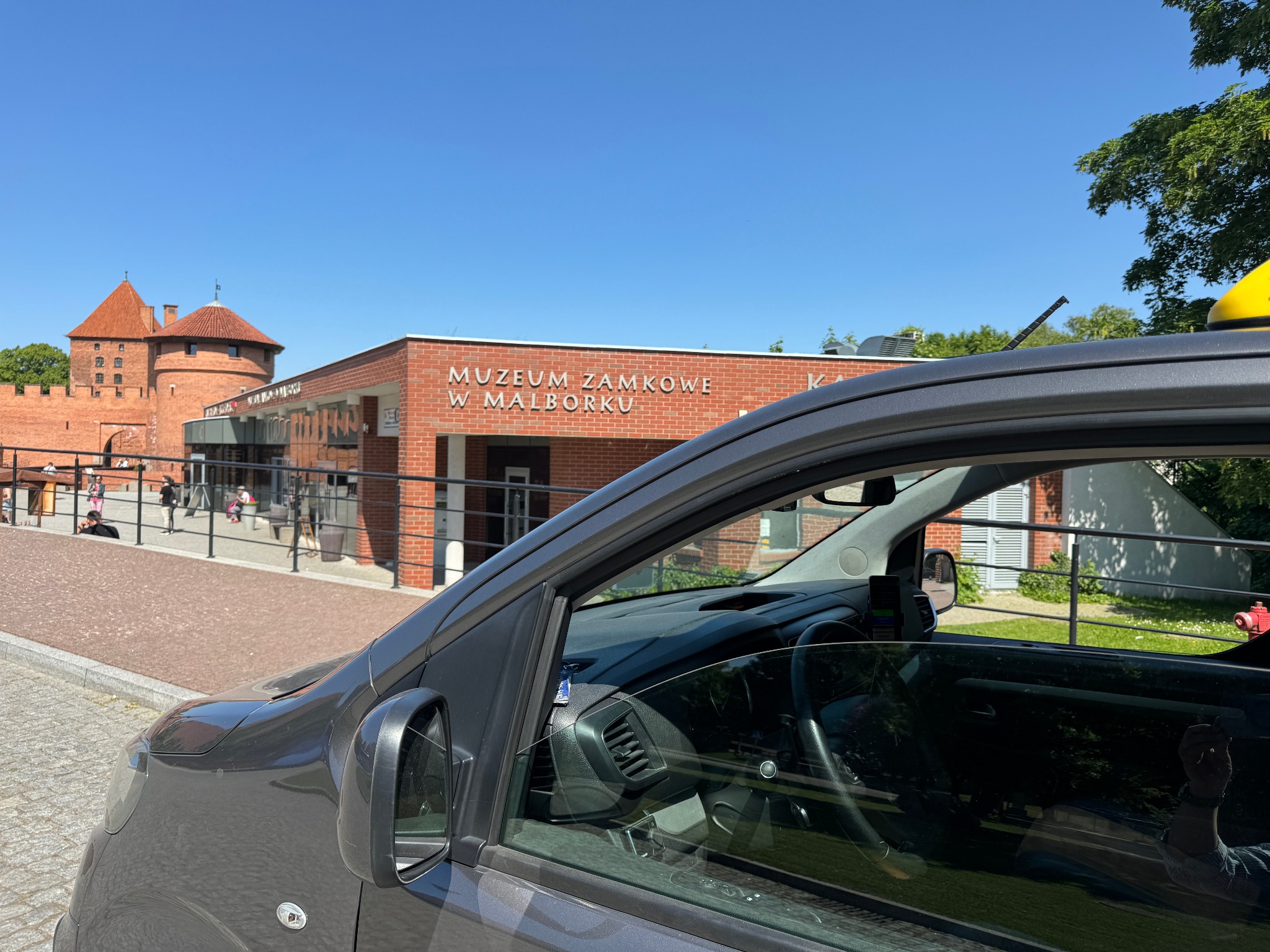 Szary van z żółtym taksometrem na dachu, zaparkowany przed Muzeum Zamkowym w Malborku w słoneczny dzień.
