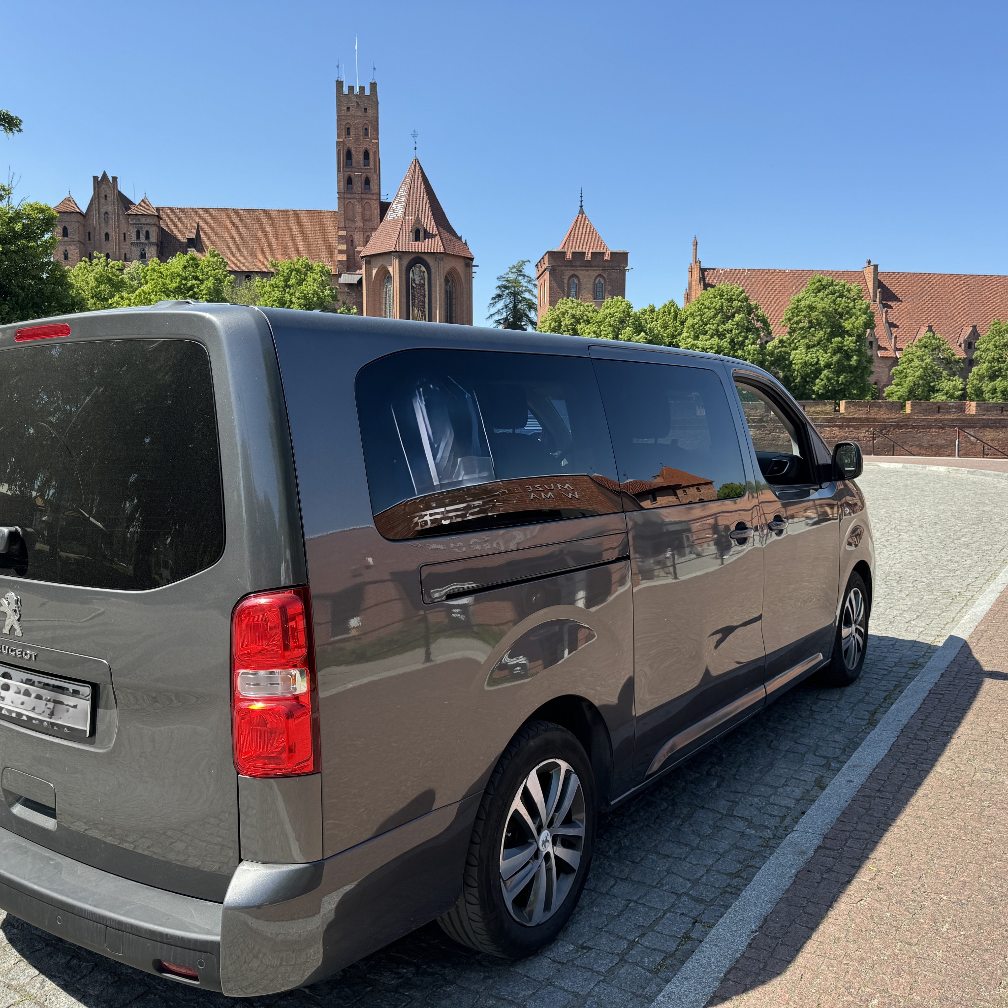 Szary bus Peugeot zaparkowany na brukowanej ulicy z widokiem na Zamek Krzyżacki w Malborku, Polska.