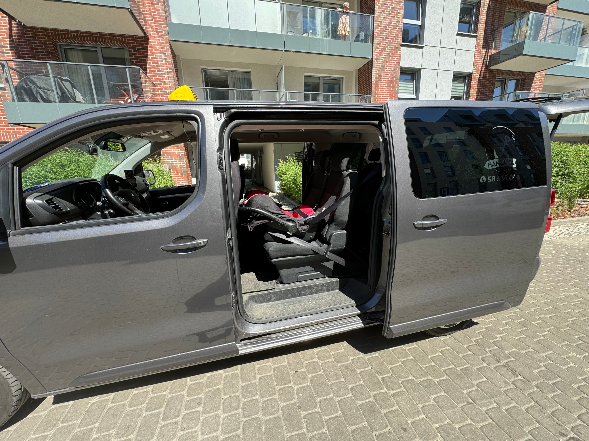 Szary van z otwartymi przesuwnymi drzwiami, widoczne foteliki dziecięce wewnątrz, taksometr na dachu, budynek mieszkalny w tle.