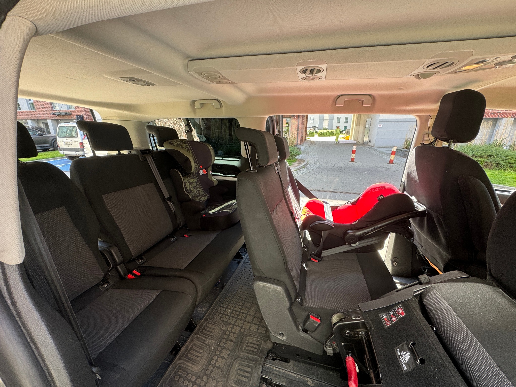 Wnętrze przestronnego busa z trzema rzędami siedzeń, wyposażonego w dwa foteliki dziecięce: jeden czerwony i jeden w kamuflażu, widok z tyłu pojazdu.