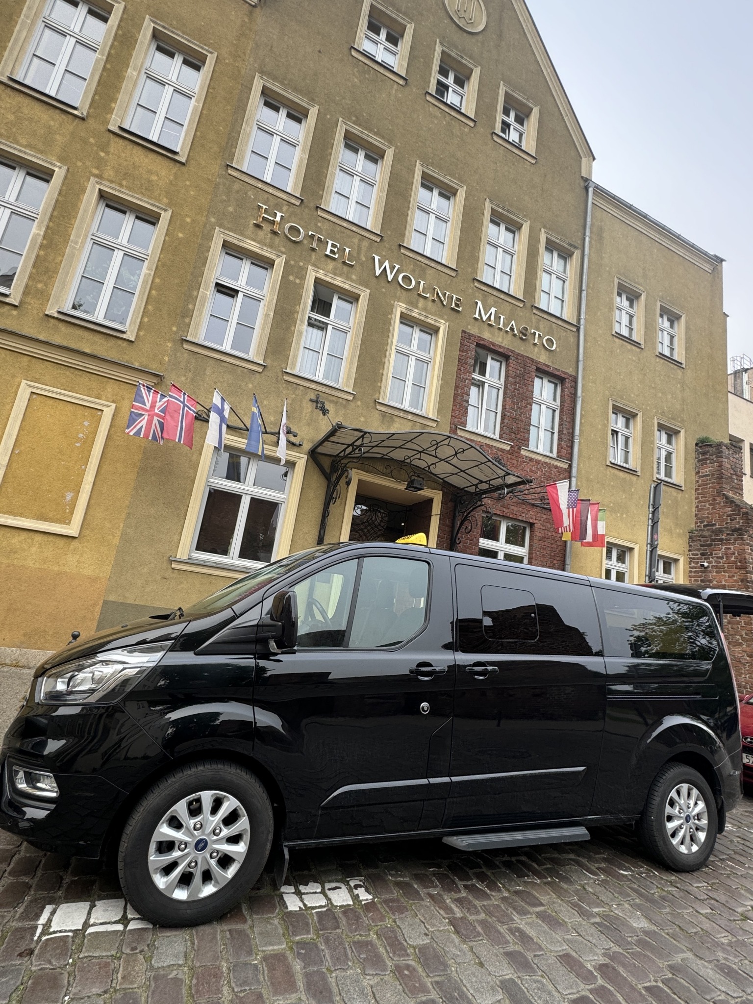 Czarny bus Ford Transit Custom z wysuwanym stopniem, zaparkowany przed Hotelem Wolne Miasto z powiewającymi flagami różnych państw.